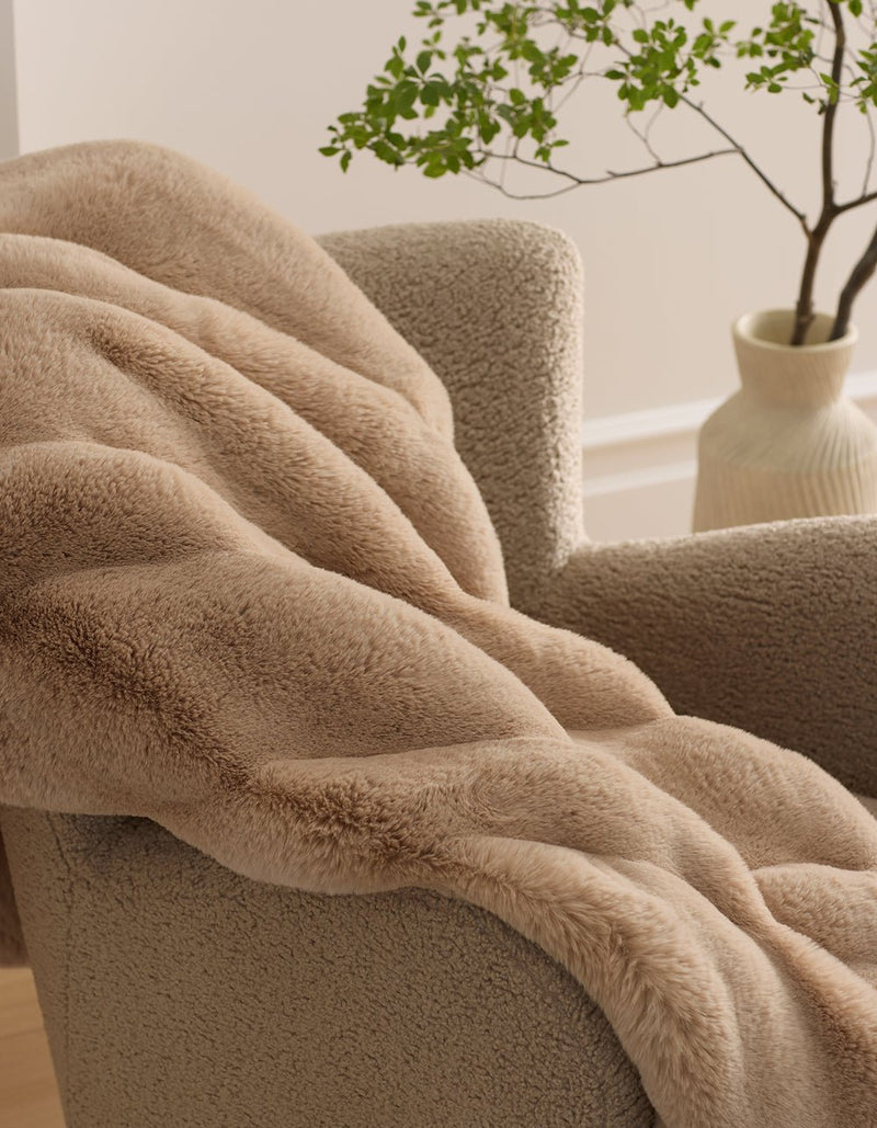 The Cozy Earth Channel Cuddle Blanket, in soft beige faux fur, is draped over a textured armchair. In the background, a light-colored vase displays green leafy branches.