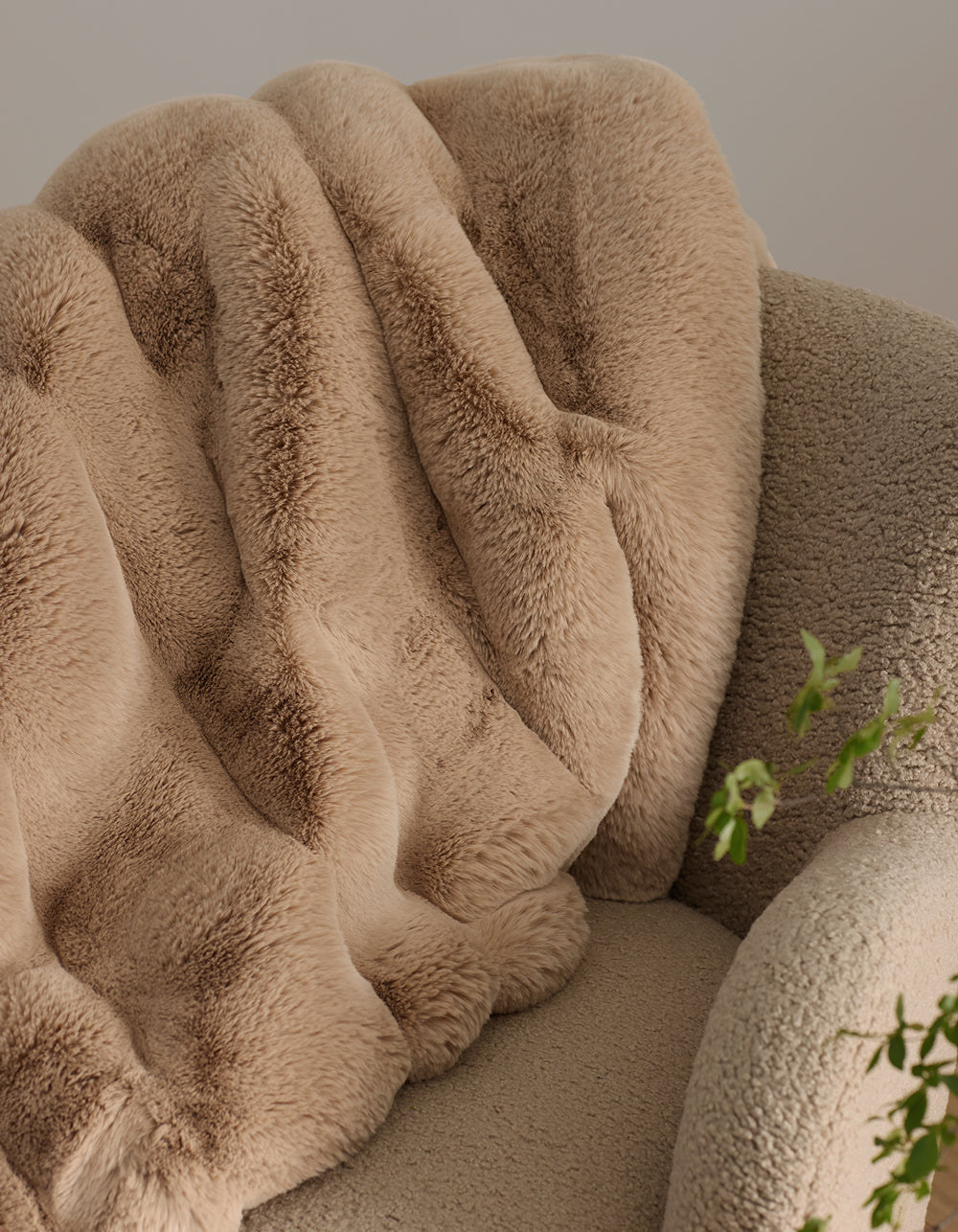 The Channel Cuddle Blanket by Cozy Earth, a plush beige faux fur throw, is draped over a textured light tan armchair. Green leaves peek in from the lower right, adding color to the inviting scene.|Color:Brandy