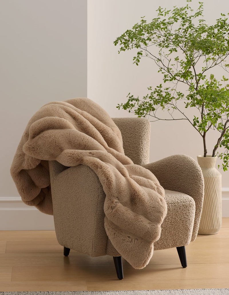 A soft, beige armchair with black legs is draped with the plush Channel Cuddle Blanket by Cozy Earth. Nearby, a leafy green plant in a textured white vase sits on a wooden floor against a light-colored wall.