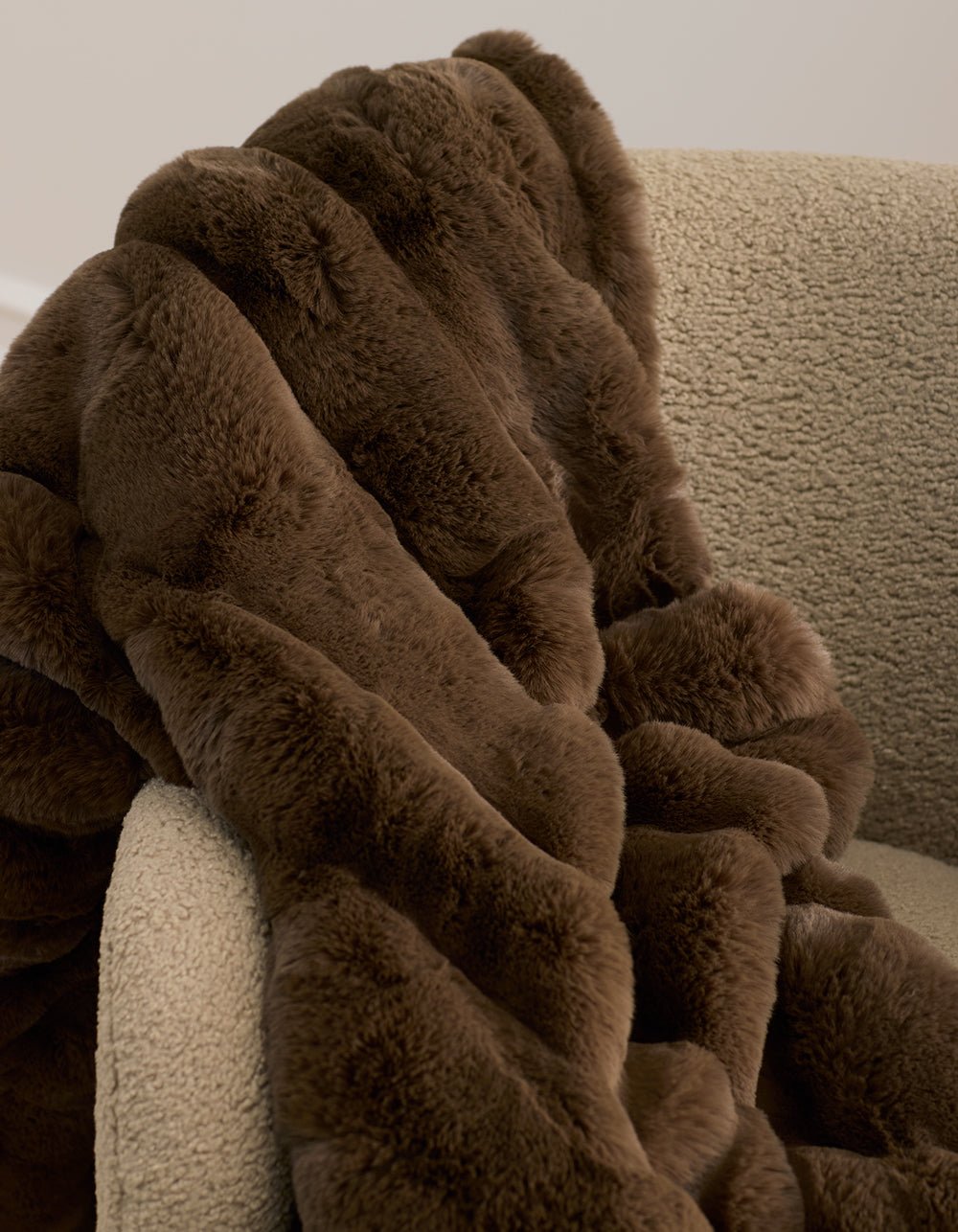 The Cozy Earth Channel Cuddle Blanket, a plush brown faux fur throw, is draped over the arm of a textured beige armchair set against a neutral background.|Color:Cedar