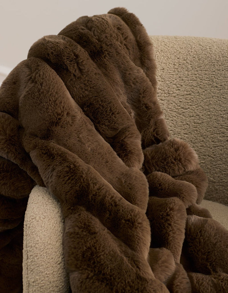 The Cozy Earth Channel Cuddle Blanket, a plush brown faux fur throw, is draped over the arm of a textured beige armchair set against a neutral background.