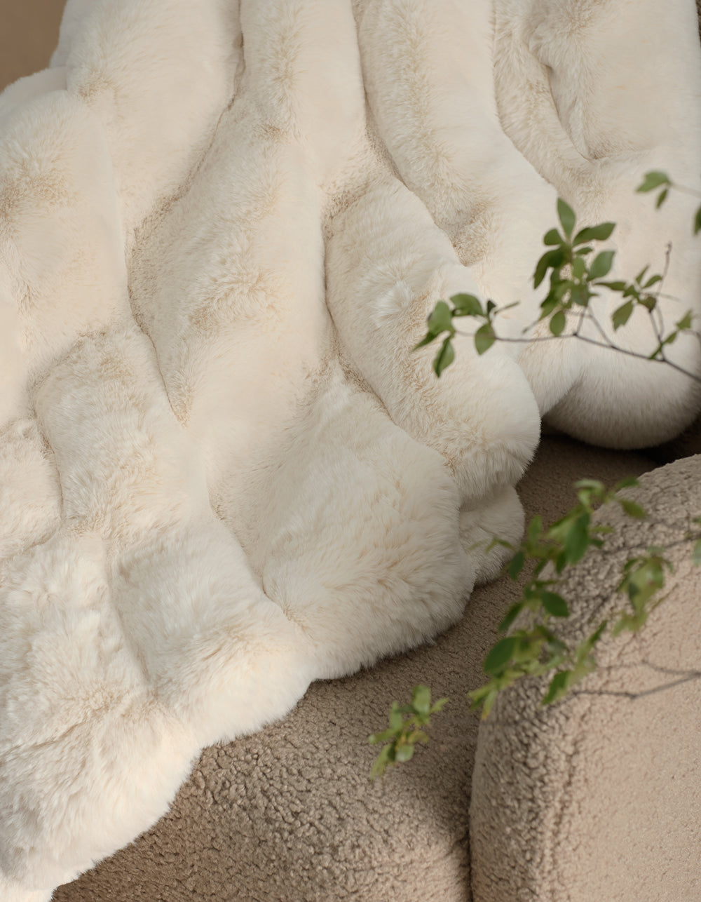 The Cozy Earth Channel Cuddle Blanket, in soft creamy white faux fur, is draped over a textured beige chair with a green leafy plant in the foreground.
