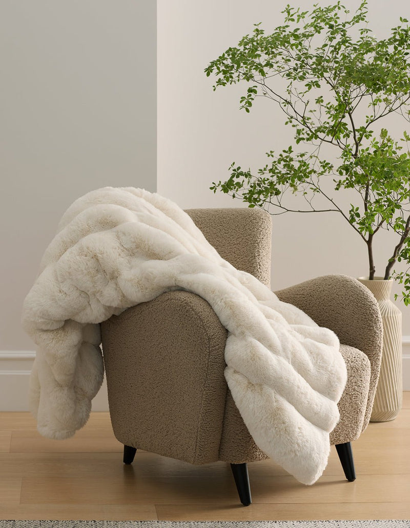 A taupe armchair with the Cozy Earth Channel Cuddle Blanket draped over it sits on a light wood floor beside a green potted plant and a light-colored wall.