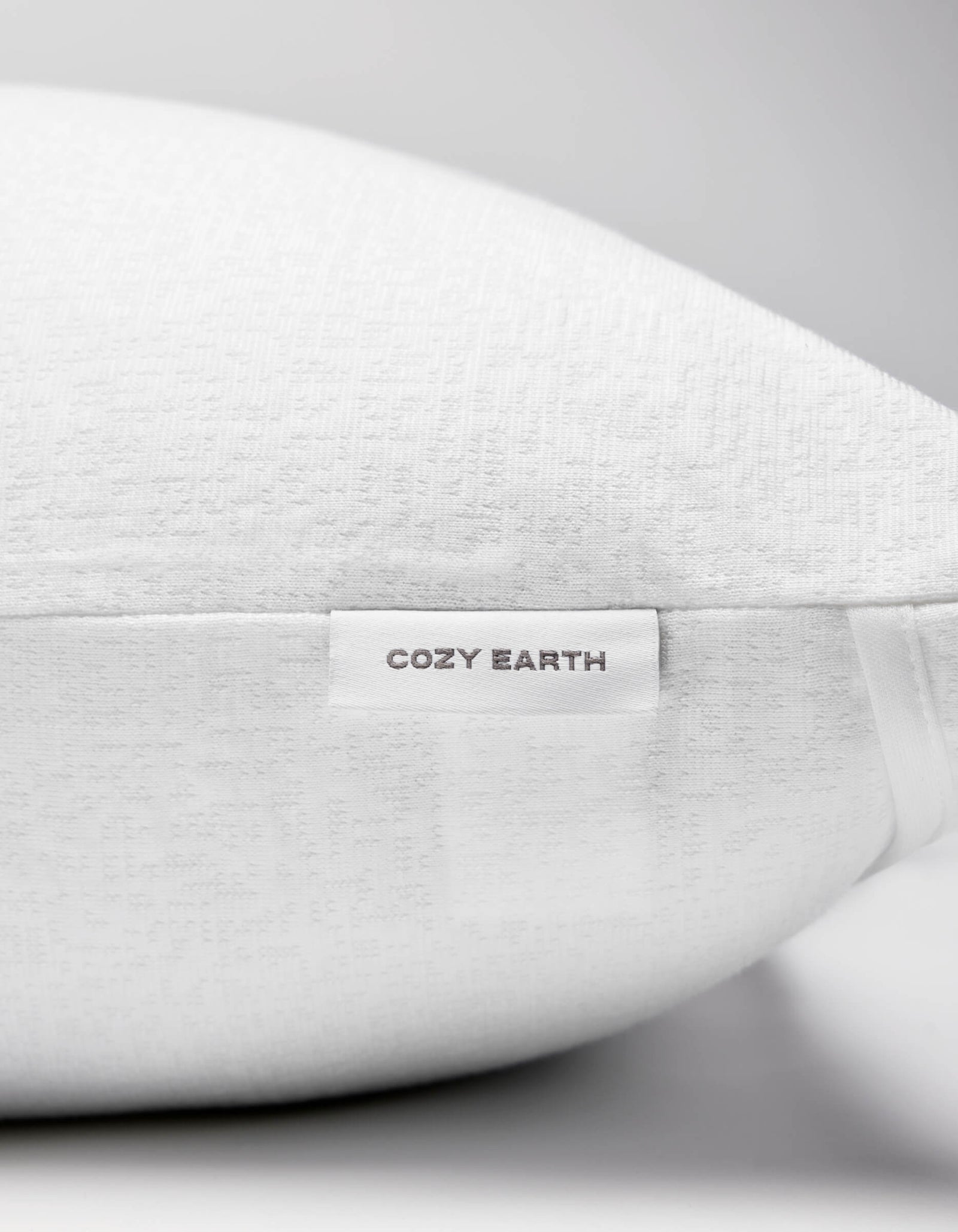 A close-up of the Cozy Earth Cooling Pillow Protector showcases its textured white fabric and a small tag reading "COZY EARTH" in gray. The all-white background creates a clean, minimalist aesthetic.