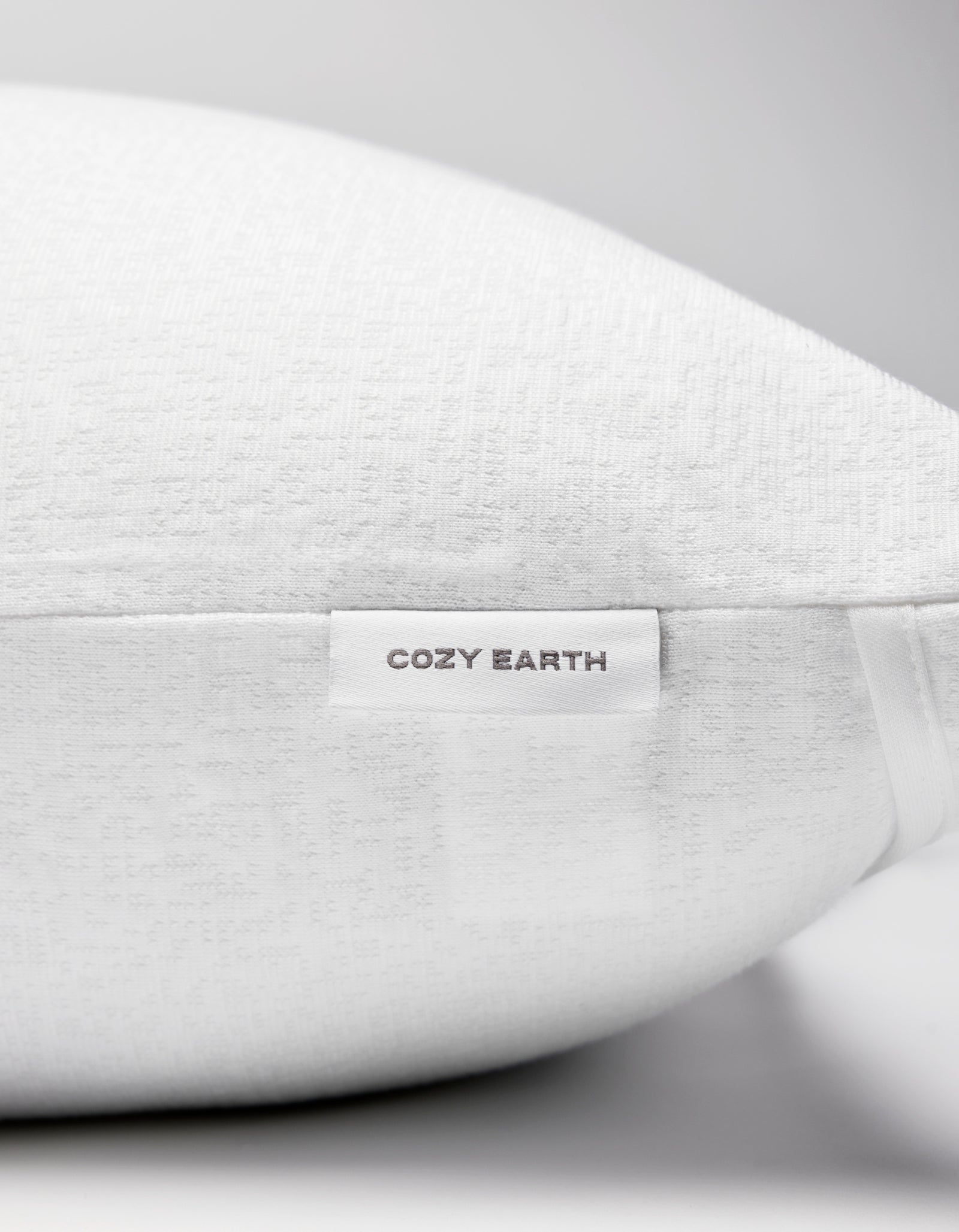 Close-up of the Cozy Earth Cooling Pillow Protector with textured white fabric and a small "Cozy Earth" label on the side. The background is plain and light-colored.