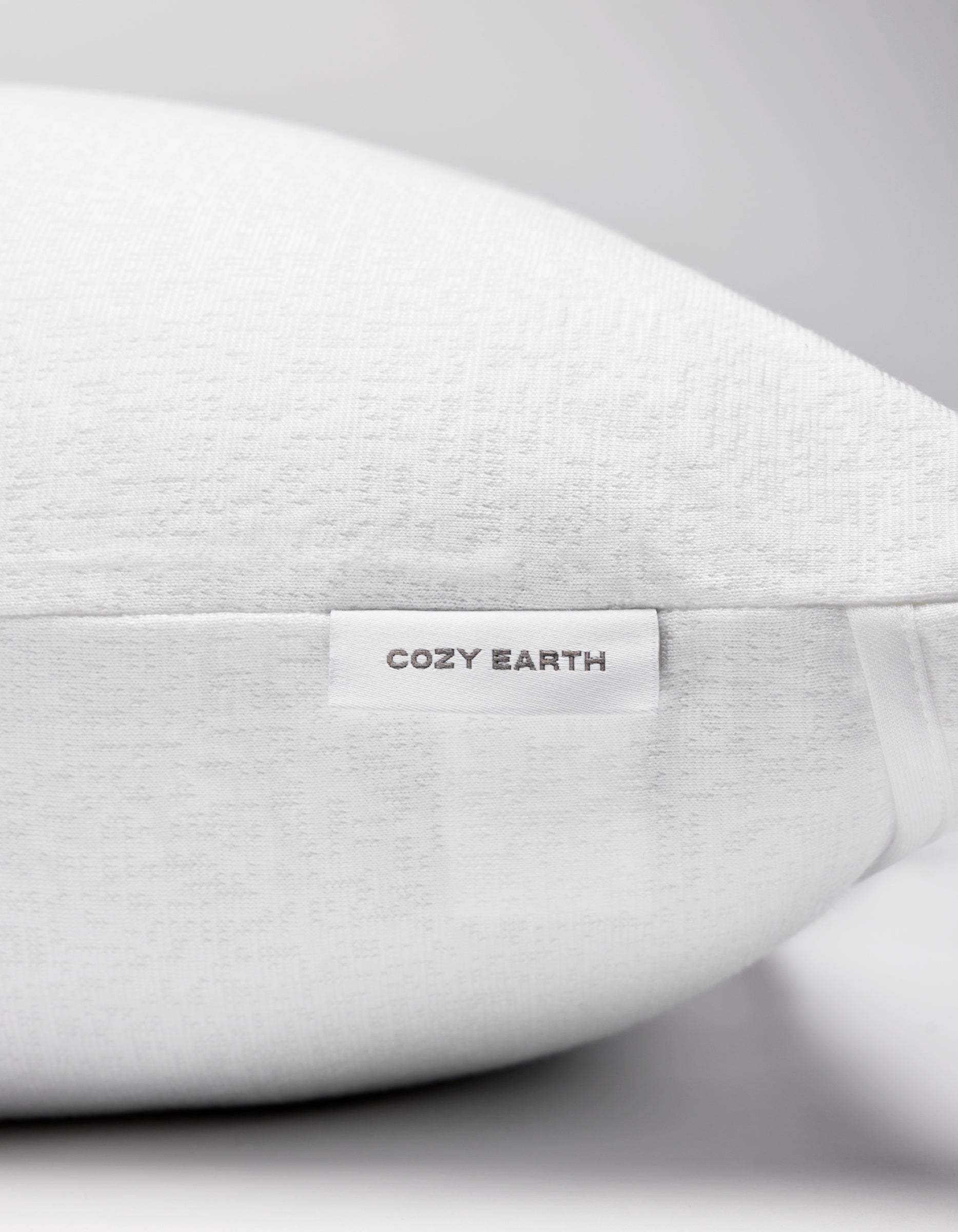 Close-up of the Cozy Earth Cooling Pillow Protector with textured white fabric and a small "Cozy Earth" label on the side. The background is plain and light-colored.