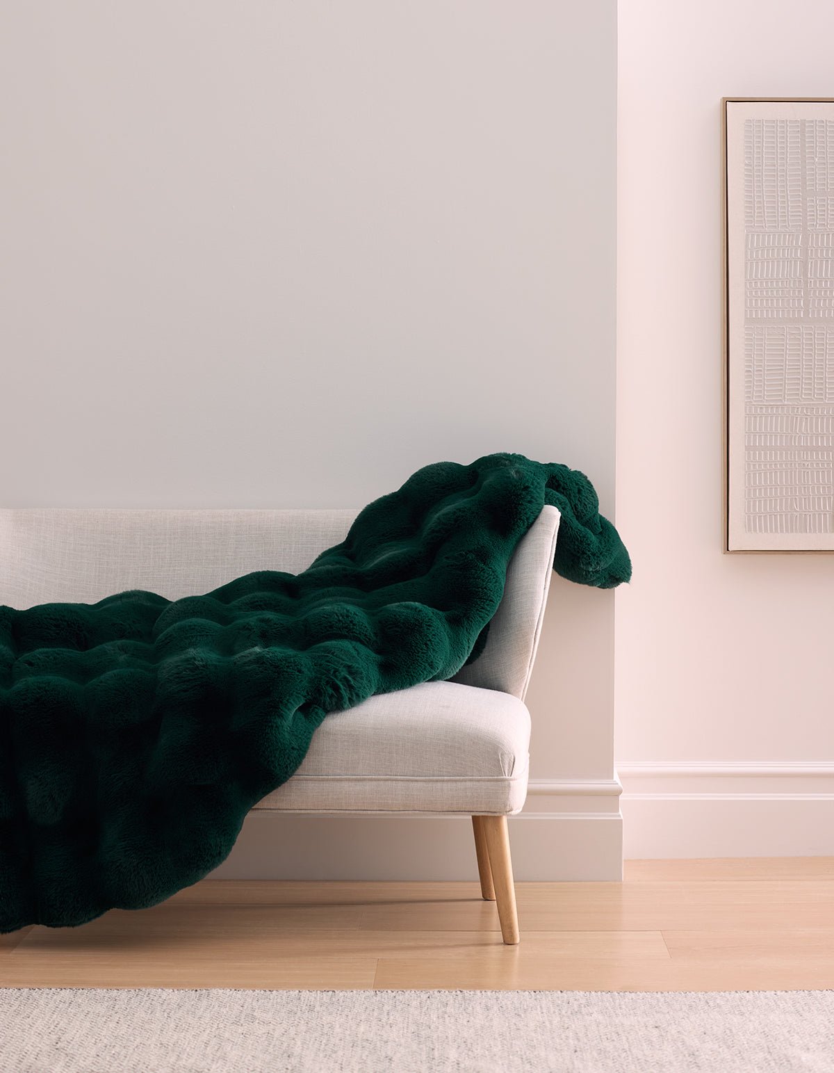 A modern, light gray bench with wooden legs is draped with the Cozy Earth Double Bubble Cuddle Blanket in dark green. The bench sits against a pale wall, beside a framed minimalist artwork on a hardwood floor.