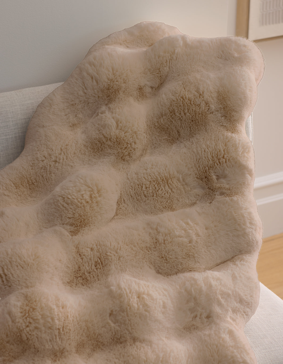The Double Bubble Cuddle Blanket by Cozy Earth, in soft, light beige faux fur, drapes over a light-colored sofa, adding warmth and comfort to a cozy, softly lit room.
