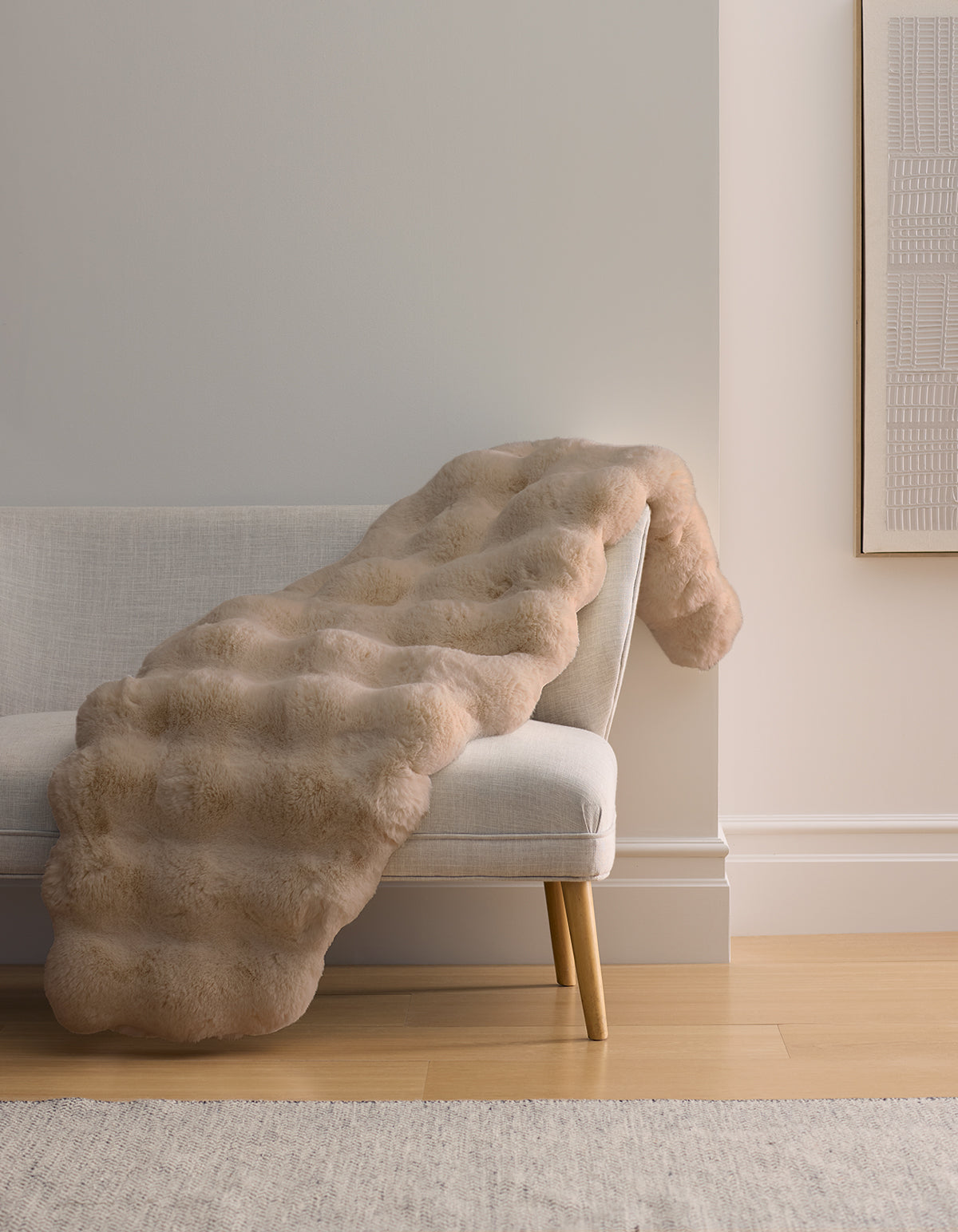 The Cozy Earth Double Bubble Cuddle Blanket, a light beige faux fur throw with a wavy quilted texture, is draped over the arm and seat of a modern light gray sofa with wooden legs in a minimalist living room.