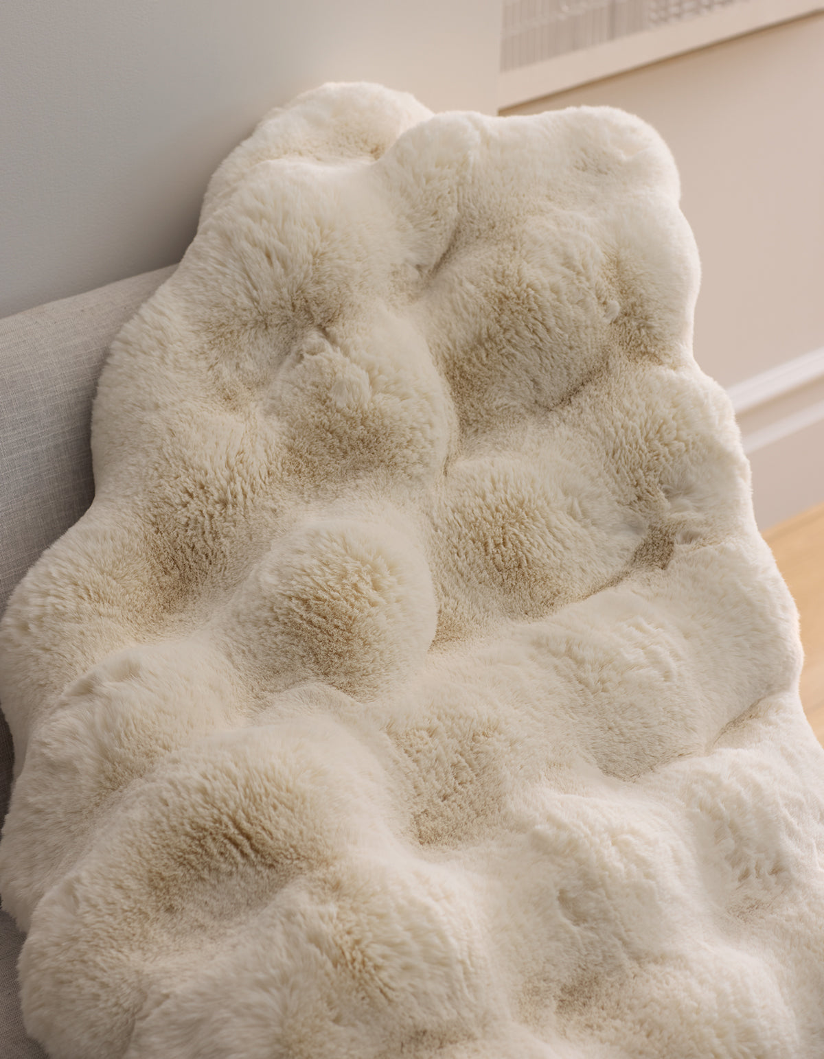 The Cozy Earth Double Bubble Cuddle Blanket, featuring a cream-colored faux fur with a textured, tufted design, is draped over a light chair in a stylish, comfortable room.