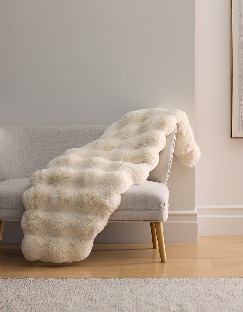 The Cozy Earth Double Bubble Cuddle Blanket in cream drapes over a light gray bench with wooden legs, set against a neutral wall and hardwood floor.