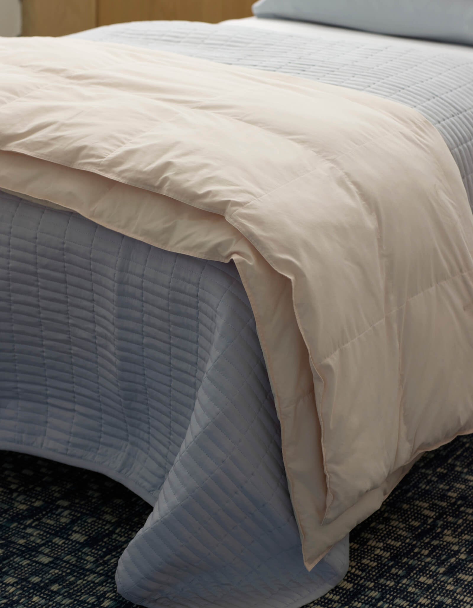 A close-up of a neatly made bed featuring the Cozy Earth Down Alternative Throw, a light blue quilted bedspread, and a beige comforter folded at the foot, all set on a dark patterned carpet.