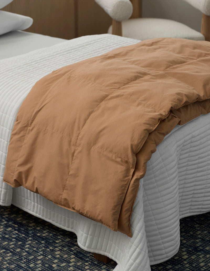 A neatly made bed features a Cozy Earth Down Alternative Throw in white, quilted and draped across the foot. In the background, a modern wooden chair with a white cushion is visible.