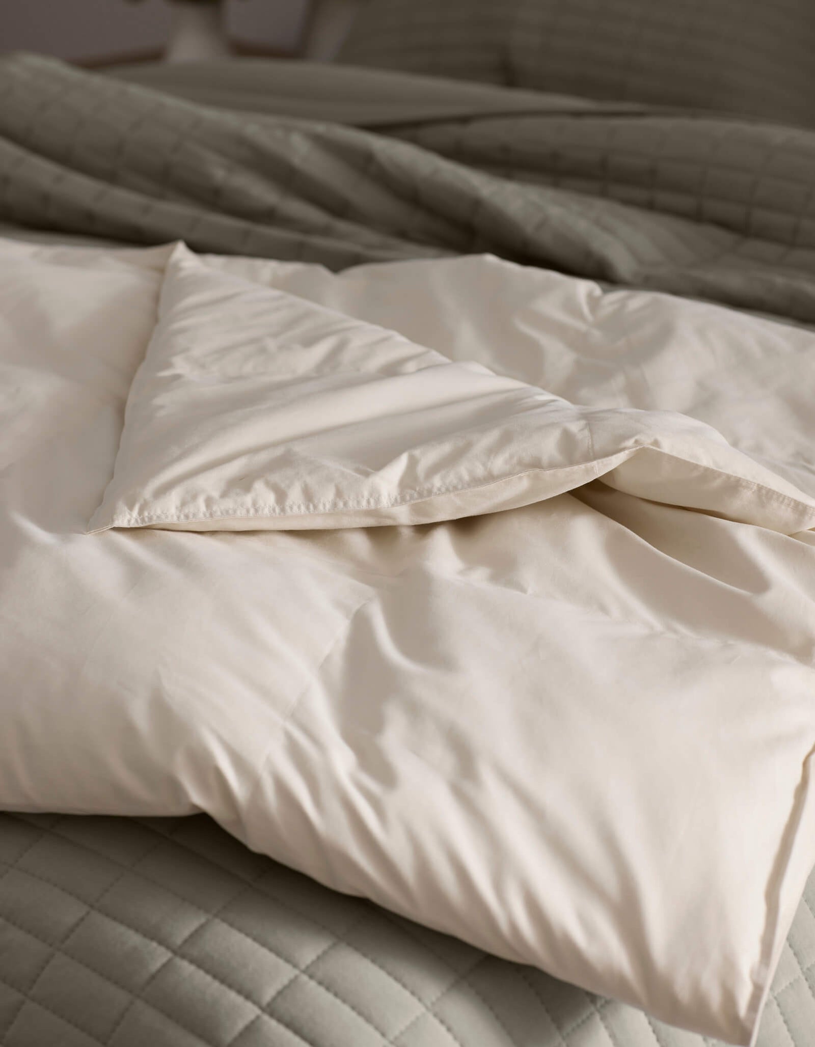 A close-up of Cozy Earth's Down Alternative Throw, folded atop a quilted light gray bedspread. The soft, cream-colored fabric looks cozy and inviting, with natural light highlighting its smooth texture.