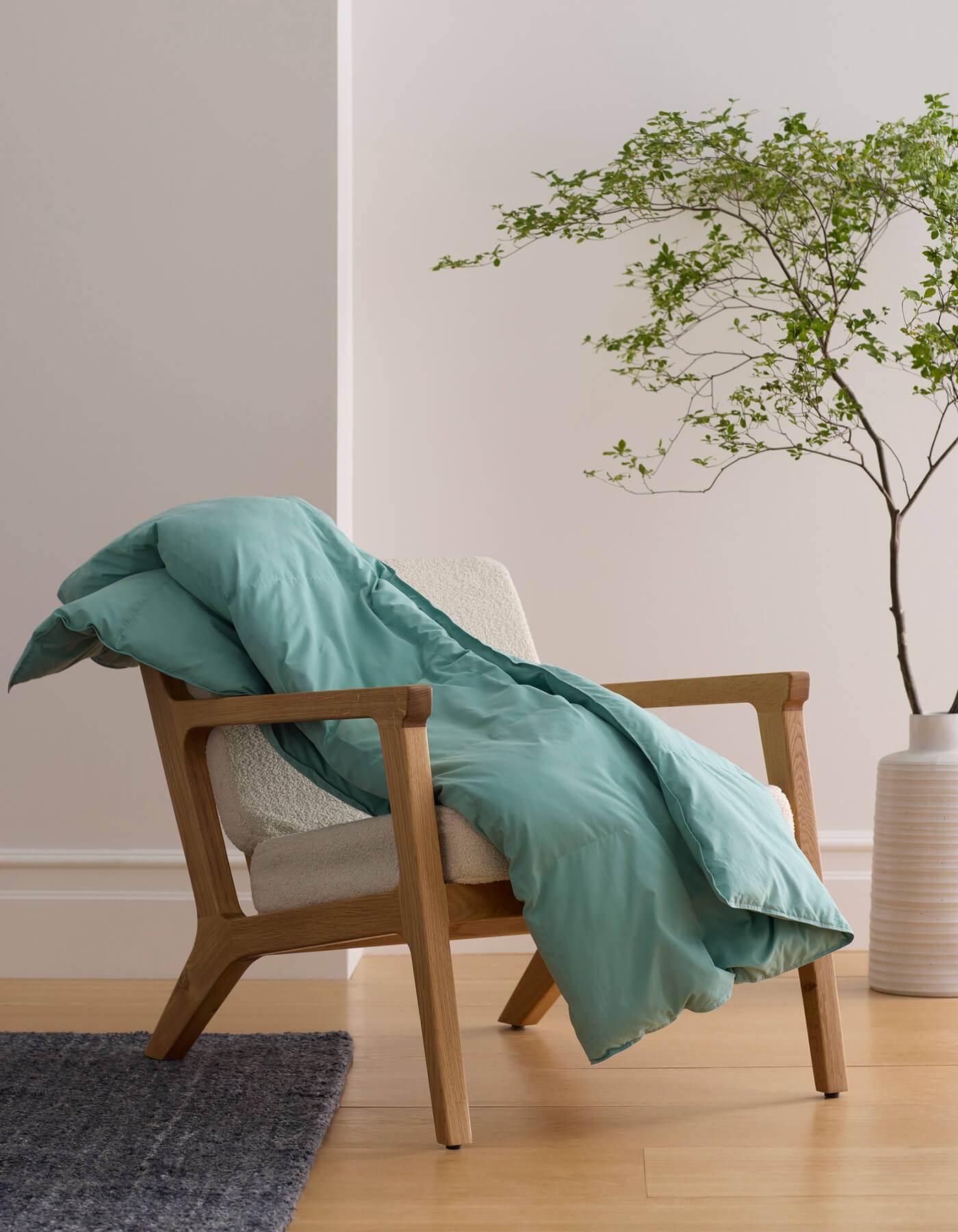 The Cozy Earth Down Alternative Throw in teal is draped over a light wood armchair with cream upholstery, beside a green leafy plant in a white vase on a wooden floor |Color:Pond