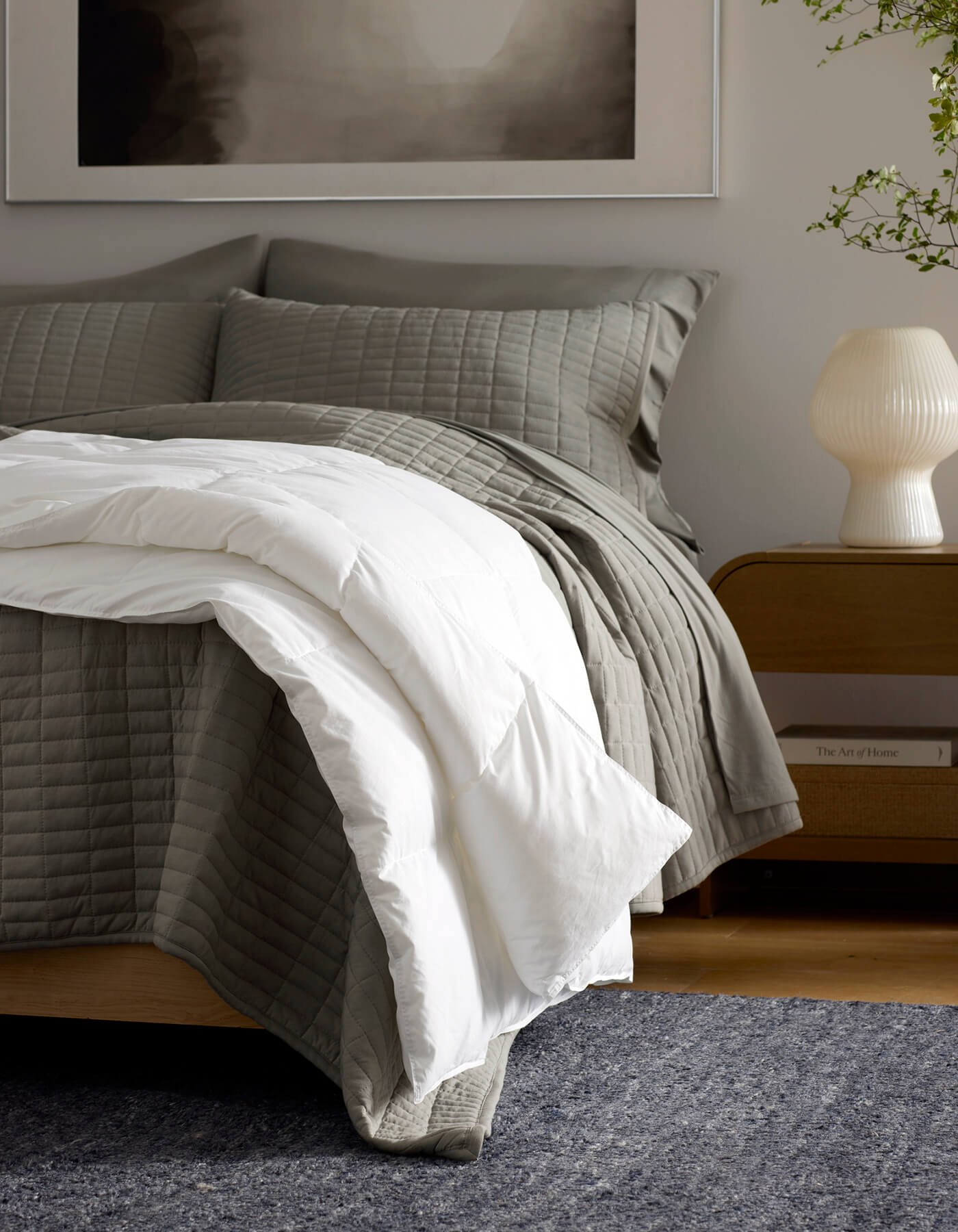 A cozy bedroom features a neatly made bed with gray quilted bedding and the Cozy Earth Down Alternative Throw, a wooden nightstand with a white lamp, and a plant in the background.