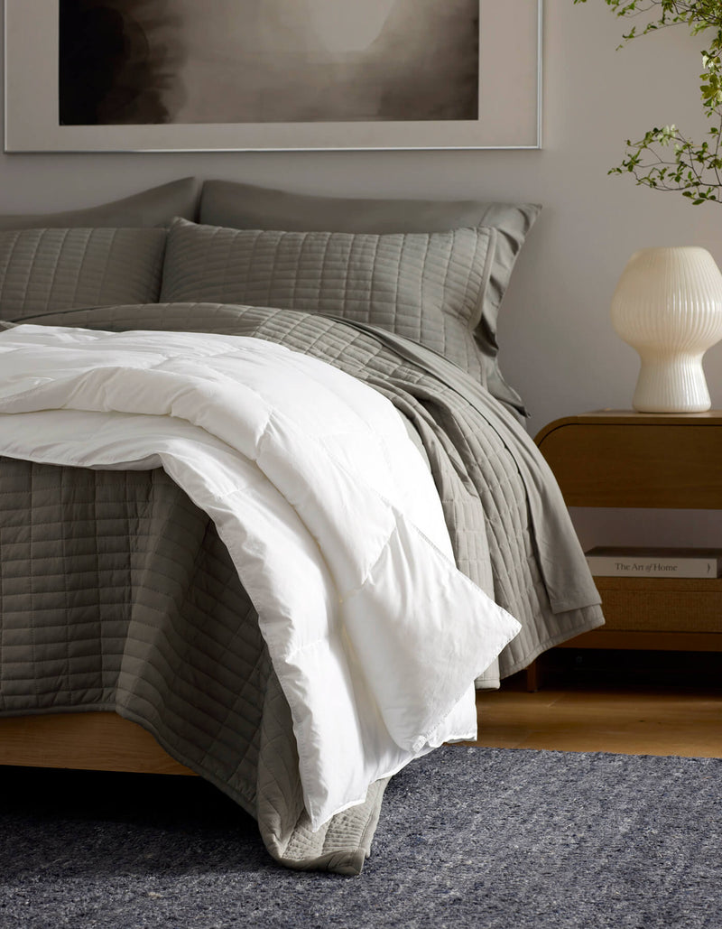 A cozy bedroom features a neatly made bed with gray quilted bedding and the Cozy Earth Down Alternative Throw, a wooden nightstand with a white lamp, and a plant in the background.