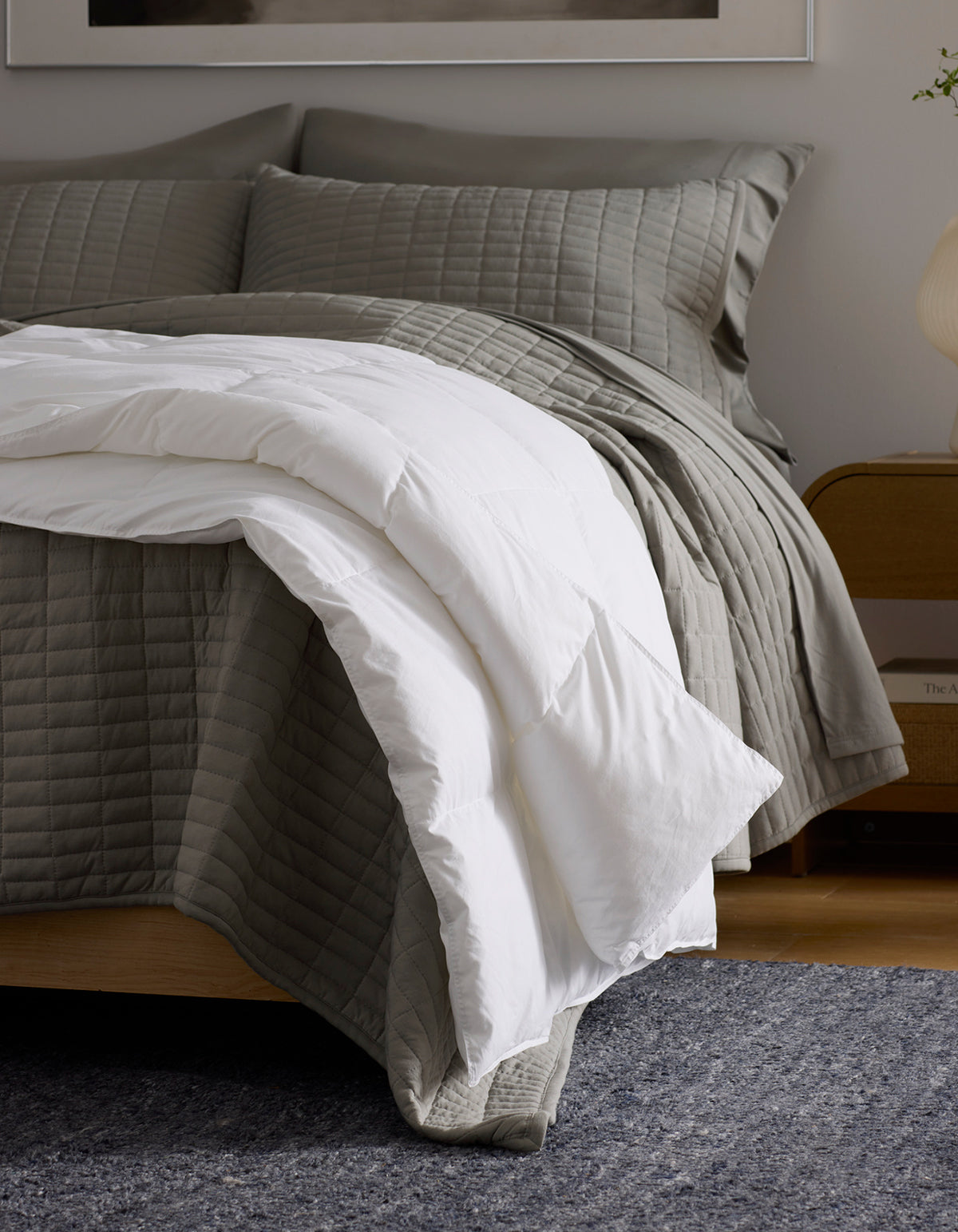 A neatly made bed with gray quilted bedding and two matching pillows is topped with a folded white Cozy Earth Down Alternative Throw. The bed has a wooden frame, sits on a dark textured carpet, and stands beside a wooden nightstand.