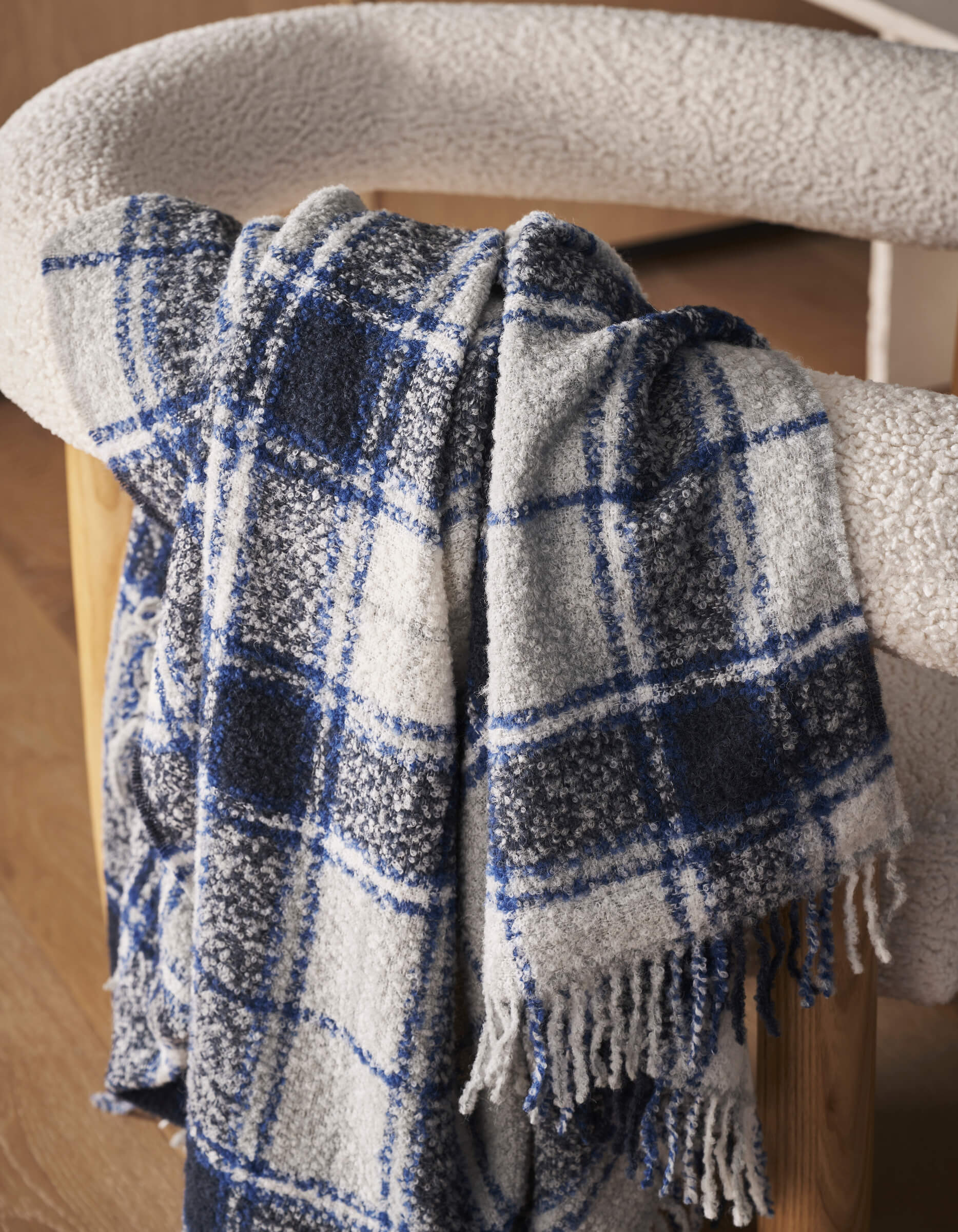 The Alpaca Boucle Throw by Cozy Earth, featuring blue, black, and white plaid with fringe, is draped over a light-textured chair in a room with warm wooden floors.|Color:Indigo Plaid
