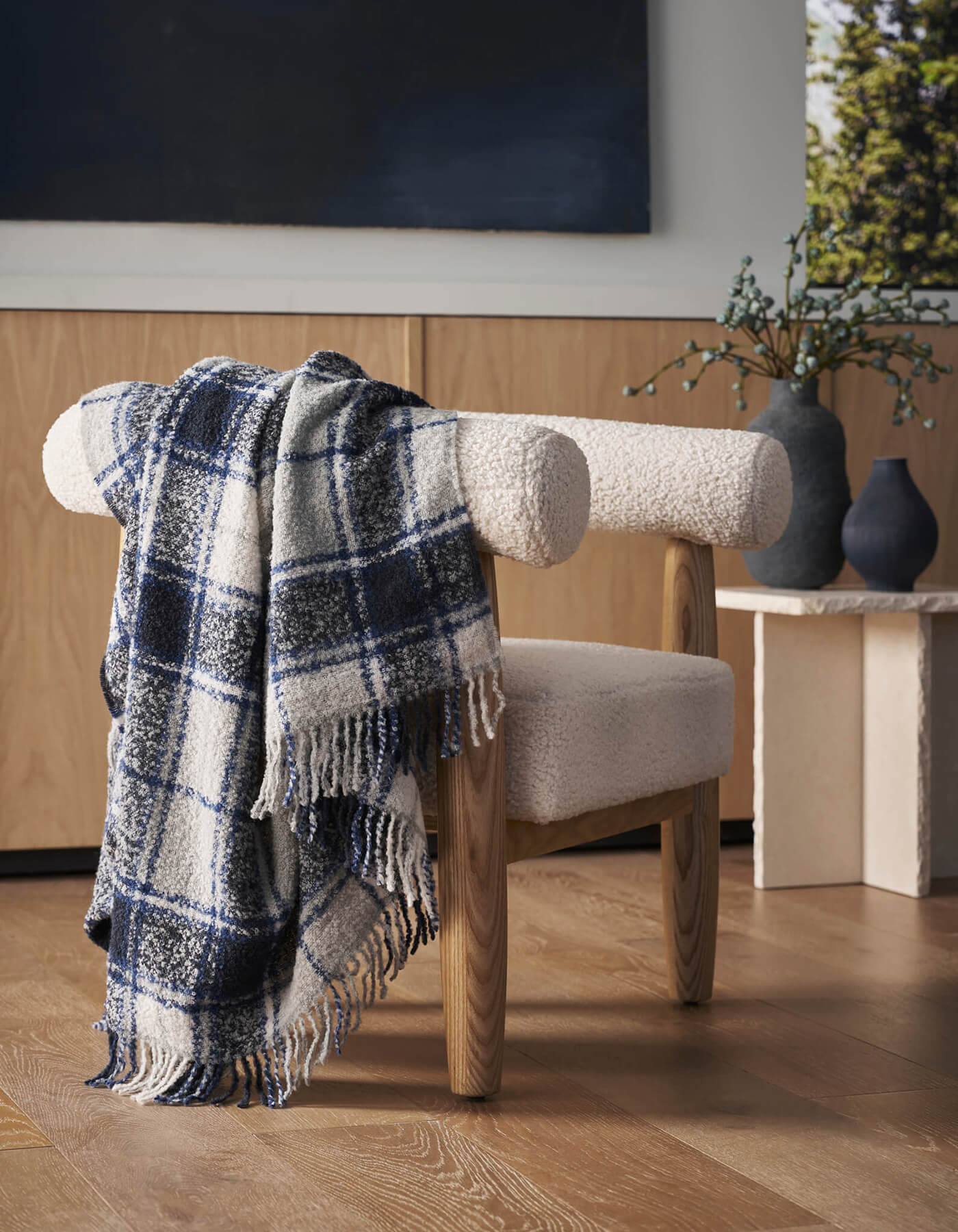 A white sherpa chair with wooden legs features the Cozy Earth Alpaca Boucle Throw draped on its back. A vase with greenery sits on a light-colored side table behind, and the setting rests on a wooden floor.