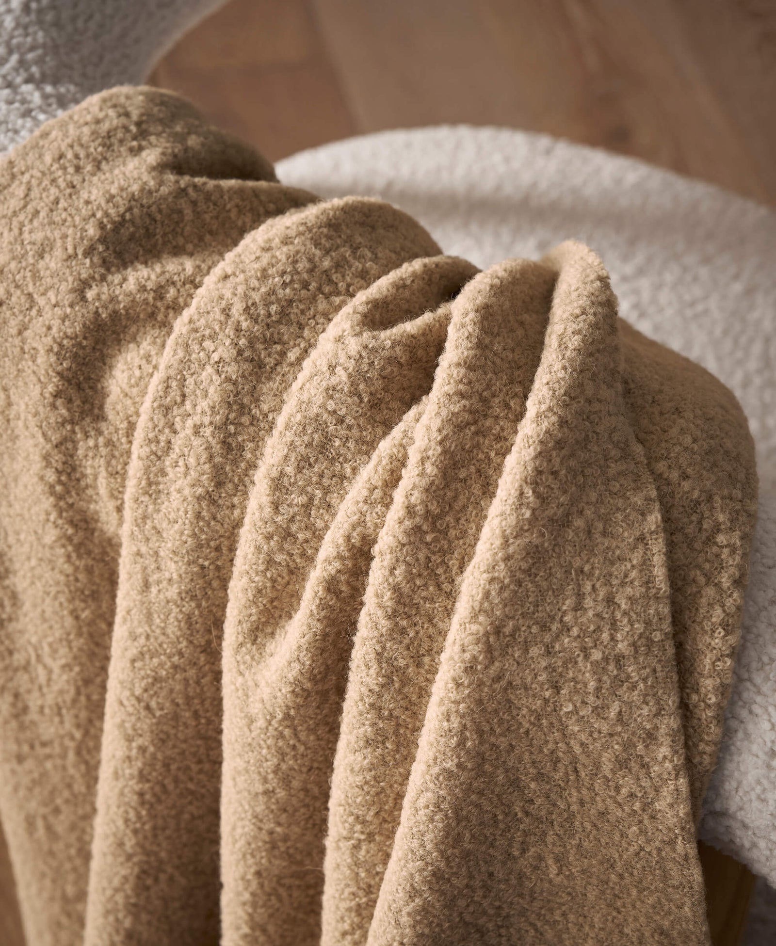 A close-up of the Cozy Earth Alpaca Boucle Throw in soft beige, elegantly draped over a white fuzzy chair, with a wooden floor in the background.