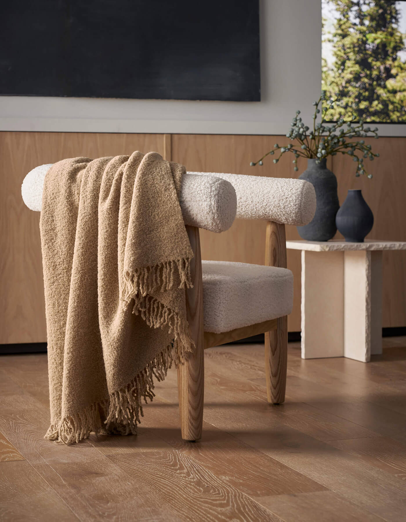 A modern wooden chair with white upholstery sits on a light wood floor, draped with the Cozy Earth Alpaca Boucle Throw. In the background, a small table displays two matte vases with greenery beside a window revealing trees outside.