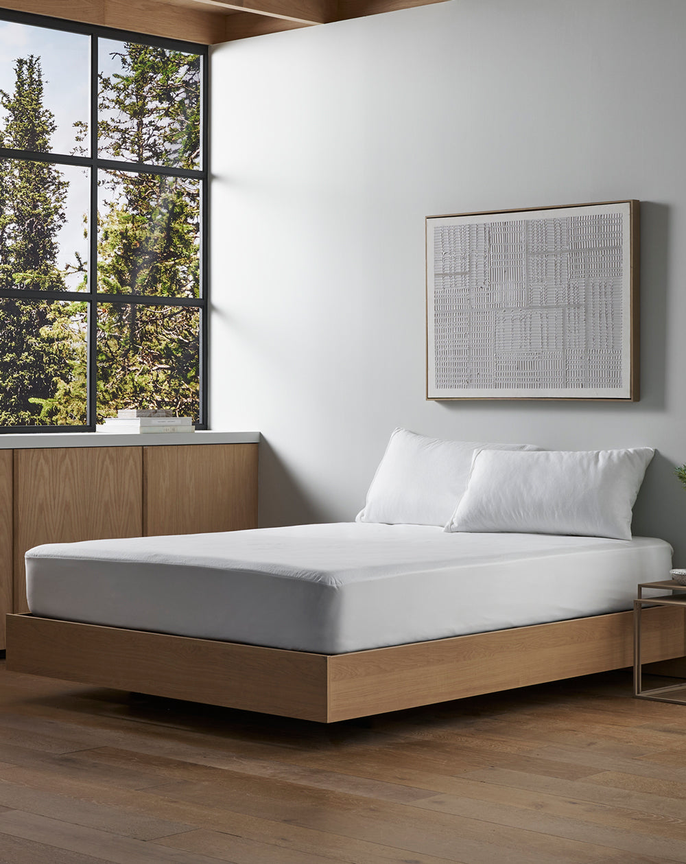 A modern bedroom features a wooden platform bed with Cozy Earth’s Cooling Mattress Protector, white bedding, light wood cabinets, a framed abstract artwork on a white wall, and a large window overlooking trees.