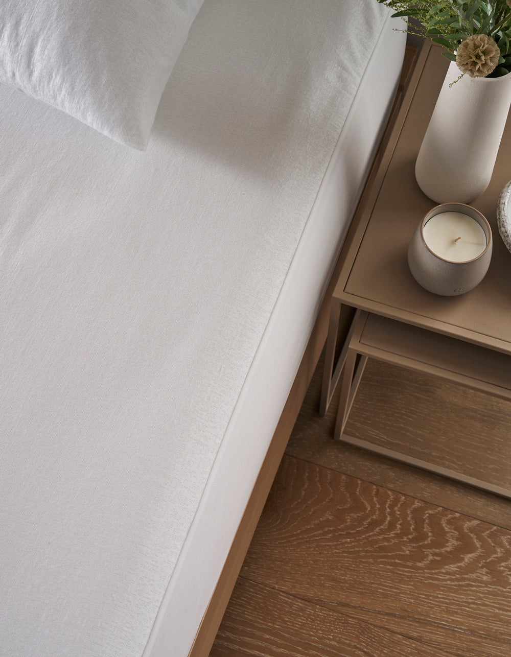 A neatly made bed with white sheets and a Cozy Earth Cooling Mattress Protector sits beside a wooden nightstand holding a lit candle, a white vase with greenery, and a stack of books on a wooden floor.