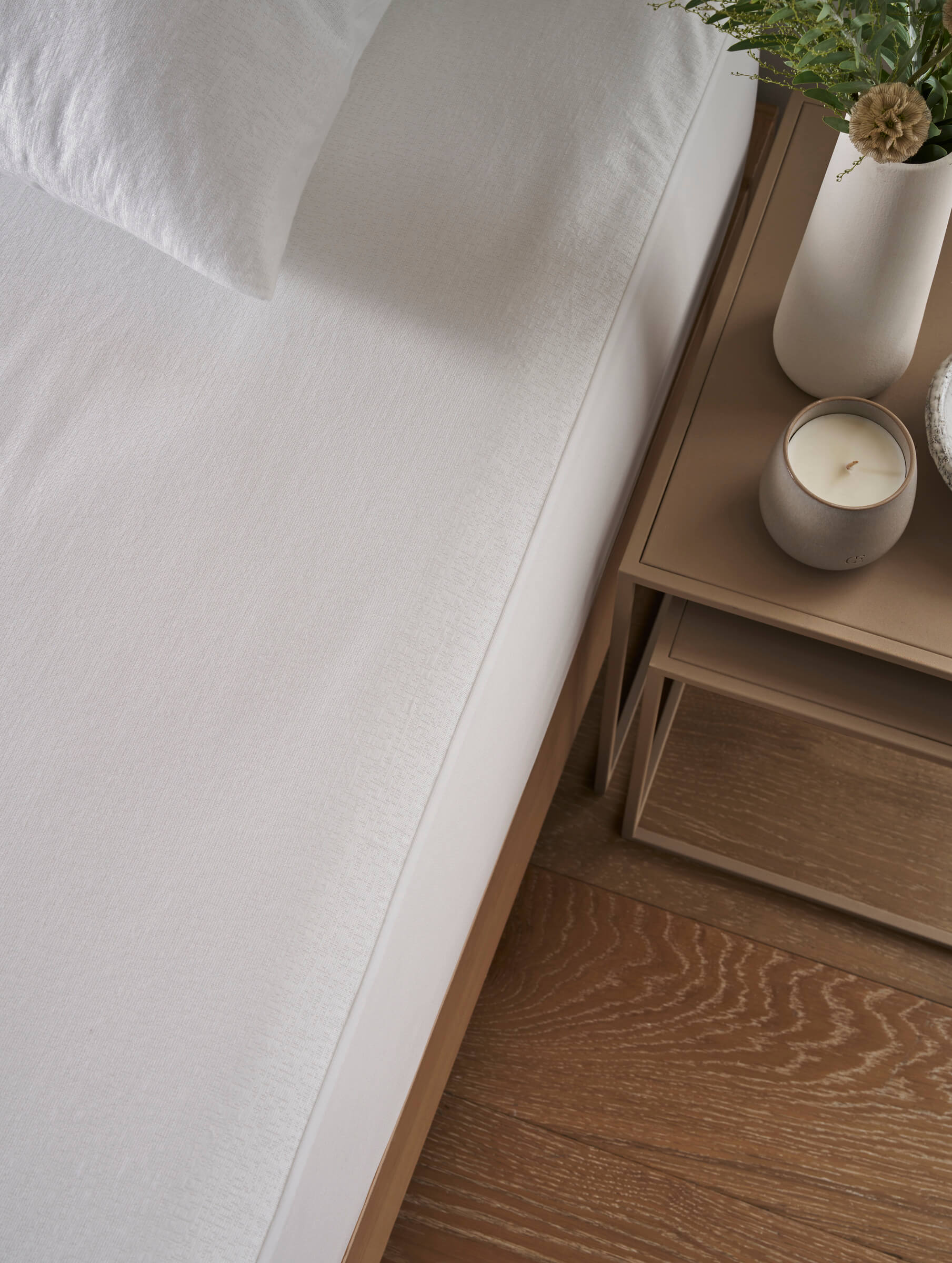 A neatly made bed with Cozy Earth's Cooling Mattress Protector is beside a wooden nightstand topped with a white vase of greenery, a closed book, and a lit candle, all set on a wooden floor.