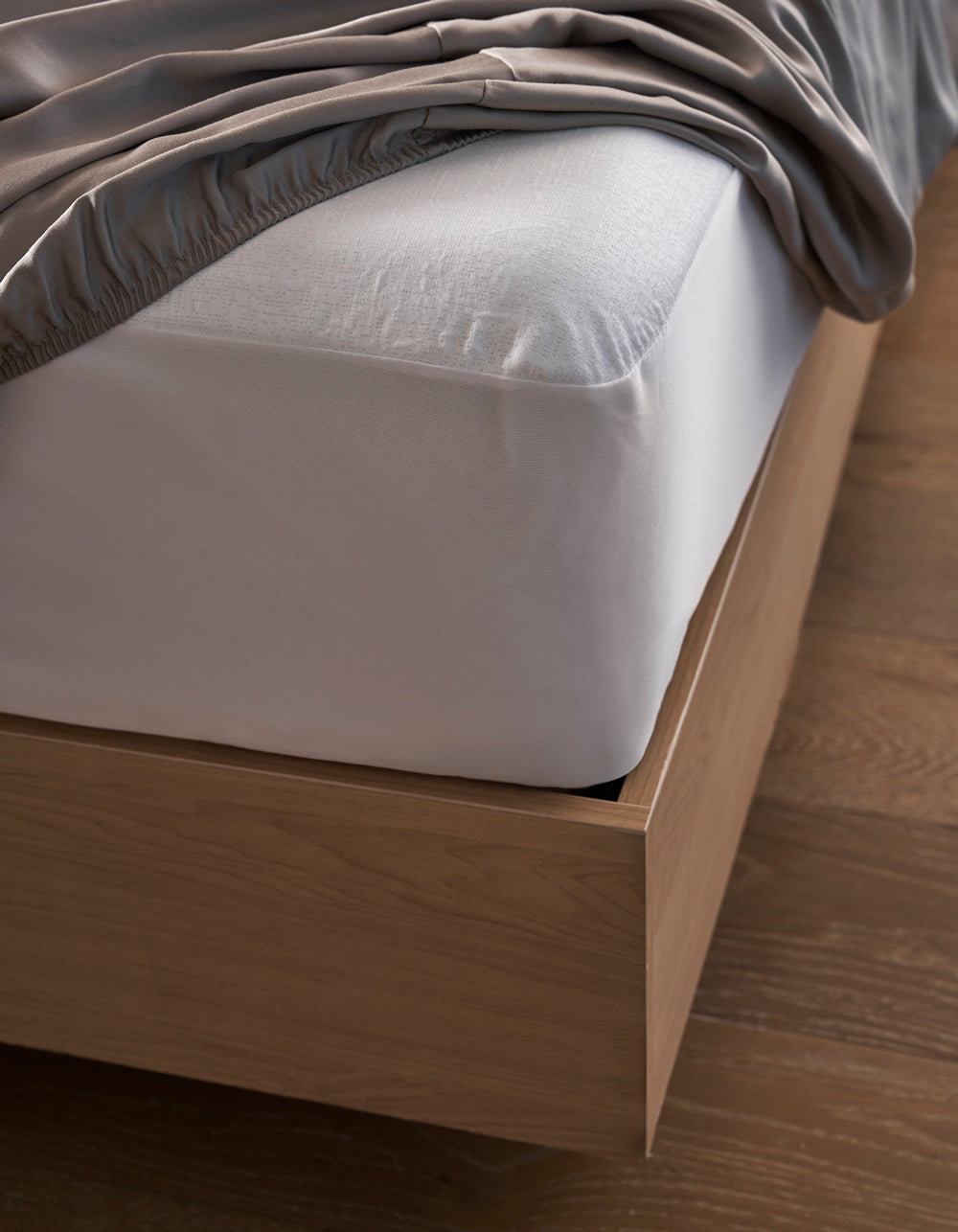 A close-up of the Cozy Earth Cooling Mattress Protector on a wooden bed frame, with a white fitted sheet and gray blanket partially draped over the mattress, set atop a wooden floor.