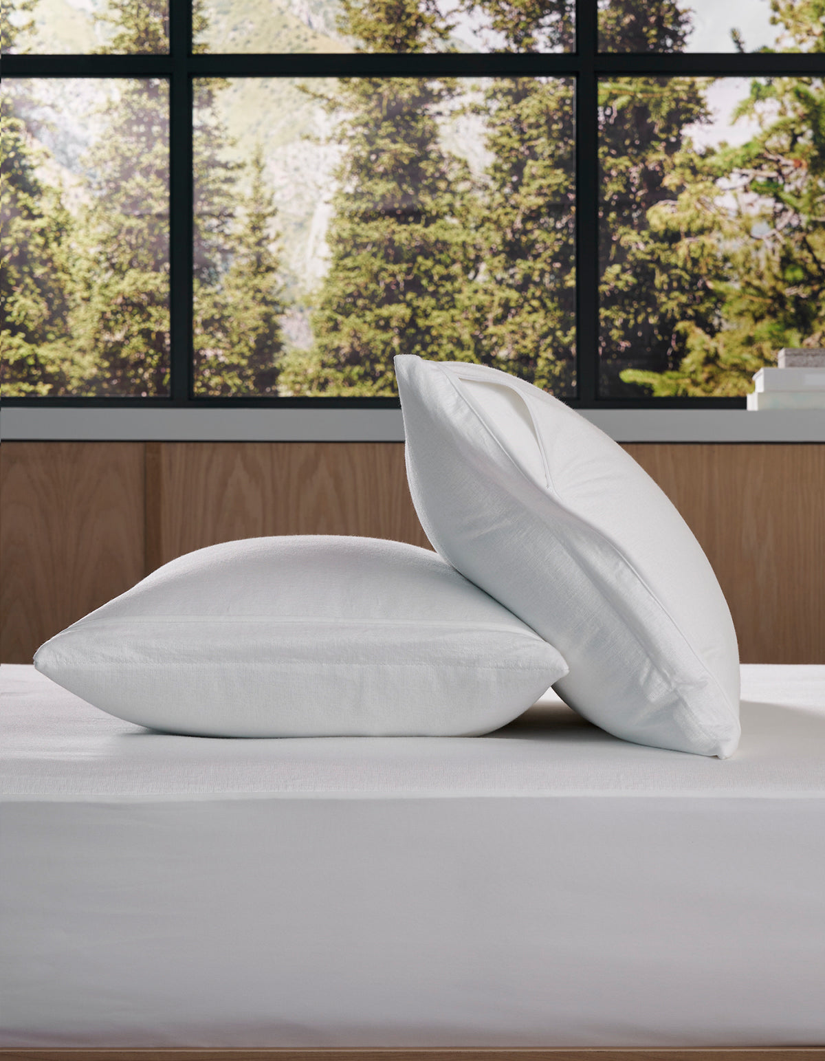 Two Cozy Earth Cooling Pillow Protectors are stacked on a neatly made bed, with a large window behind them revealing a view of green trees outside.