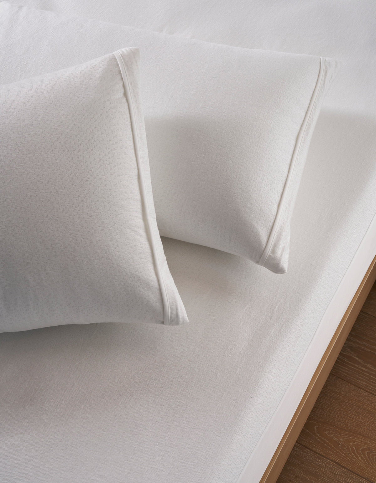 Two white Cooling Pillow Protectors by Cozy Earth lay on a neatly made bed with white sheets and a wooden frame, creating a clean, minimal, and fresh look.
