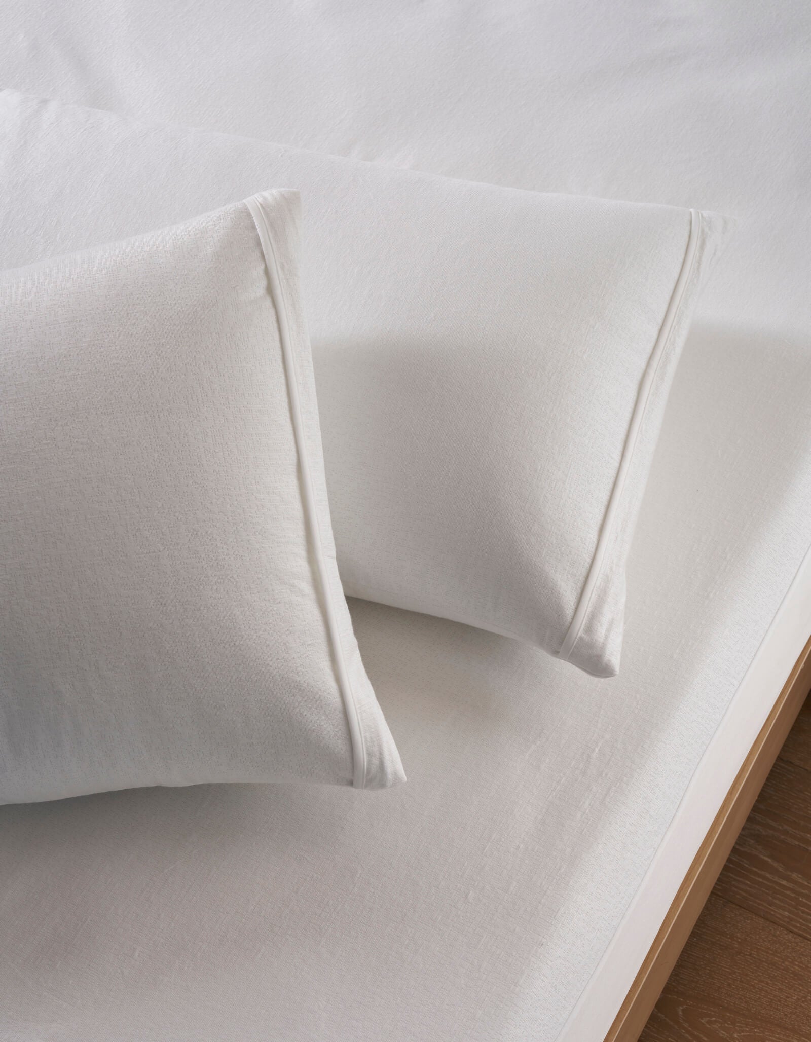 Two Cozy Earth Cooling Pillow Protectors are displayed on a neatly made bed with a white fitted sheet. The bedding appears clean and smooth, and part of the bed frame is visible in the corner.