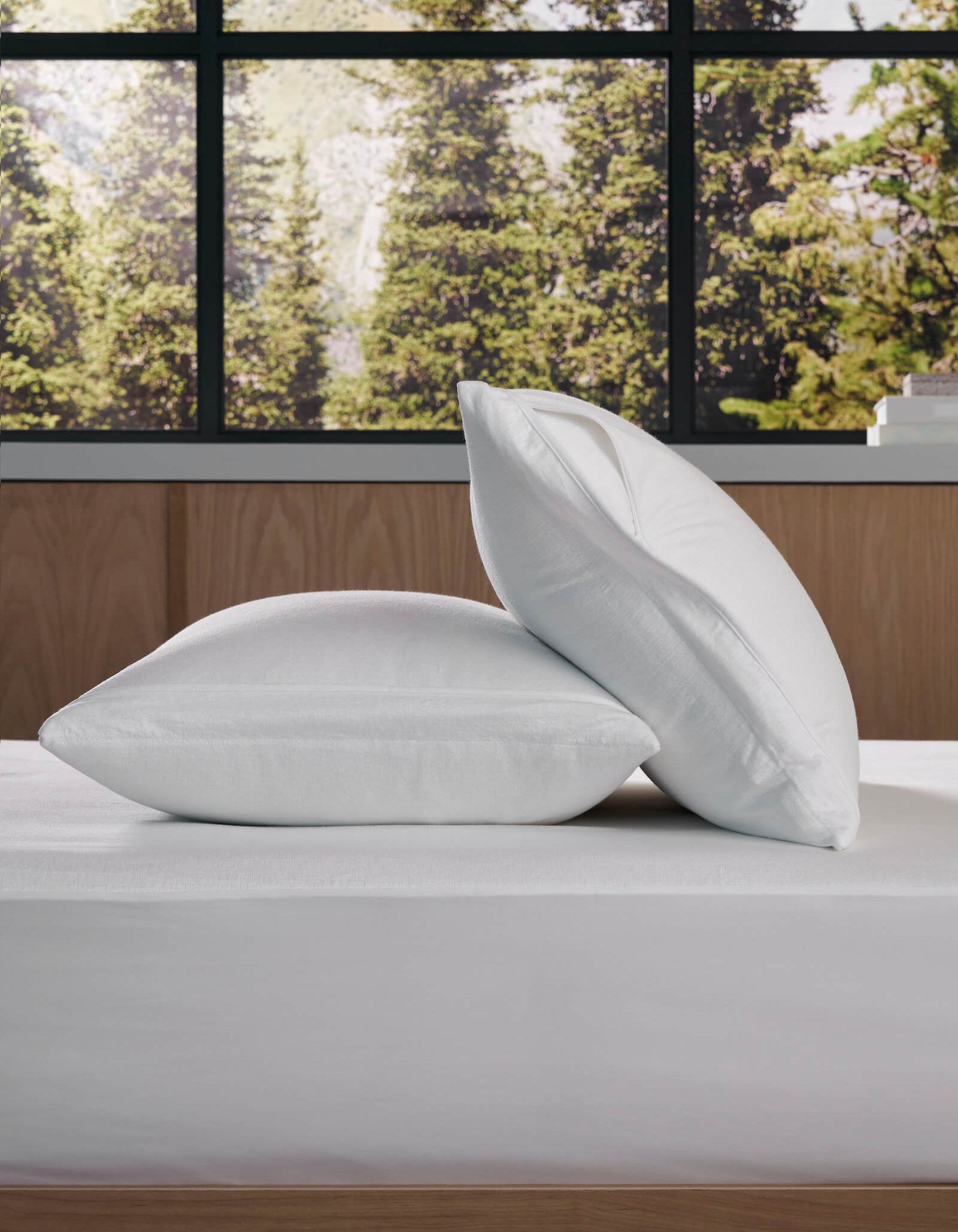 Two Cozy Earth Cooling Pillow Protectors are stacked on a neatly made bed by a large window with views of green trees, complementing the room's modern, minimalistic design.