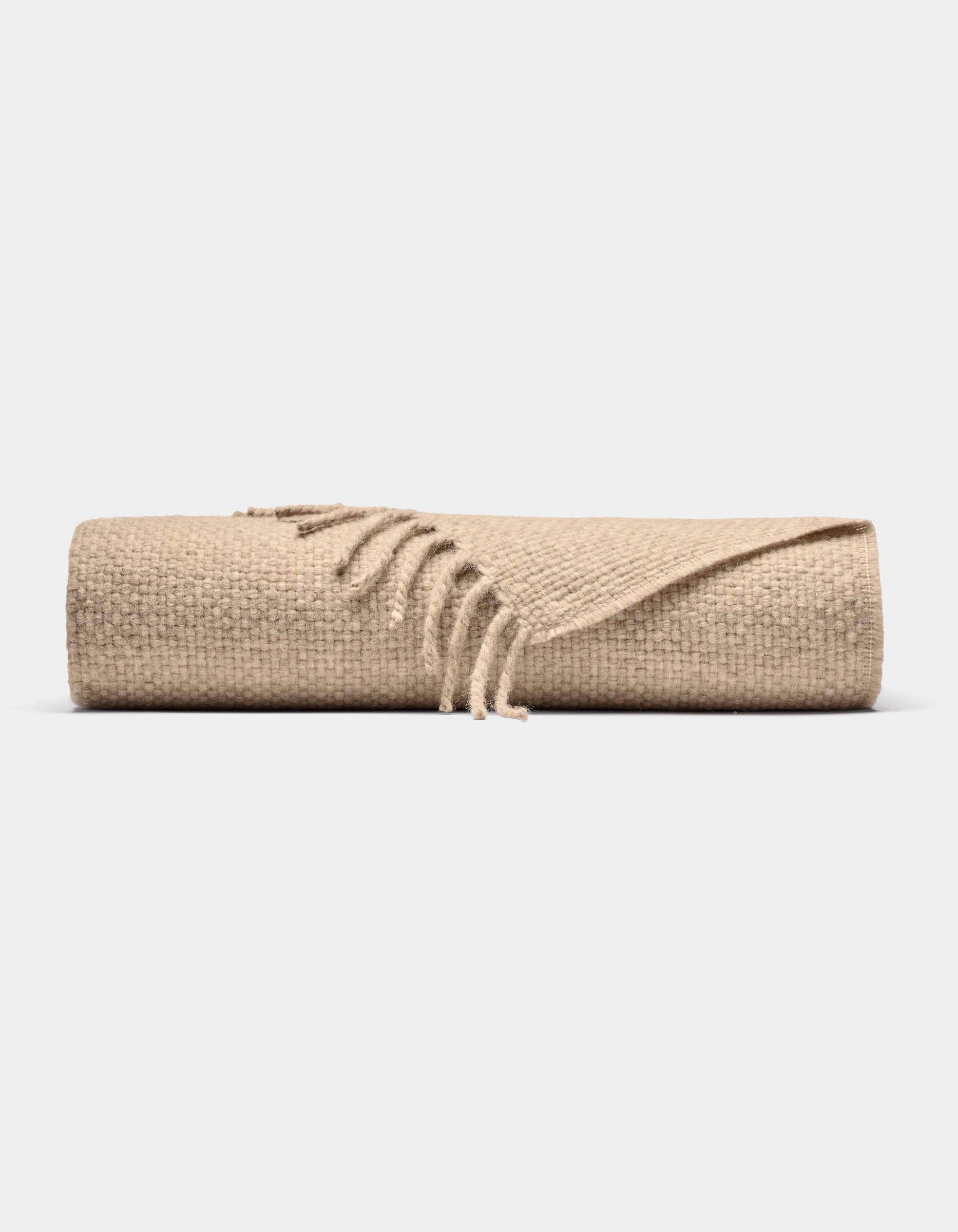 The Lucia Throw by Cozy Earth is a beige woven blanket with fringed edges, shown rolled up against a plain white background.