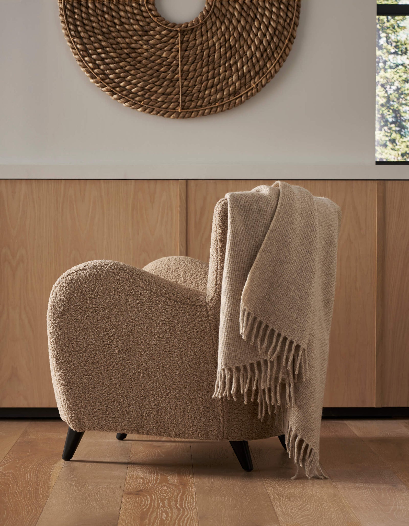 A cozy beige textured armchair with a Cozy Earth Lucia Throw draped over the back sits on a wooden floor. Behind it are light wood cabinets and a round woven wall hanging, with sunlight streaming in through the window.