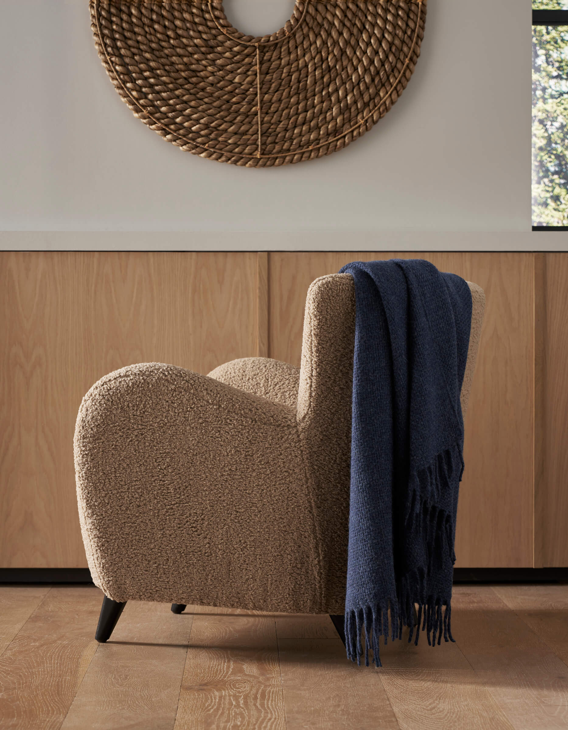 A cozy beige armchair with a Cozy Earth Lucia Throw draped over one arm sits on a wooden floor before light wood cabinets. Above the chair hangs a large, round, woven wall art piece.|Color:Indigo