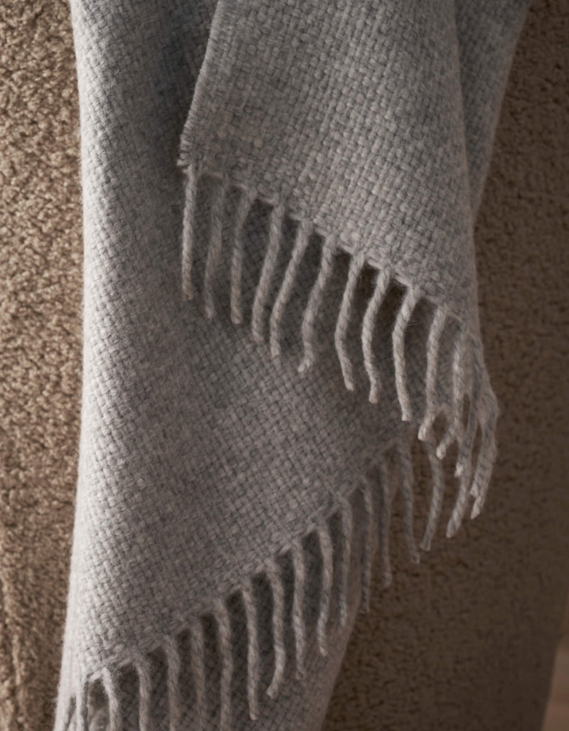 A close-up of the Lucia Throw by Cozy Earth, a soft, textured gray blanket with fringe edges, draped over a coarse tan upholstered surface.