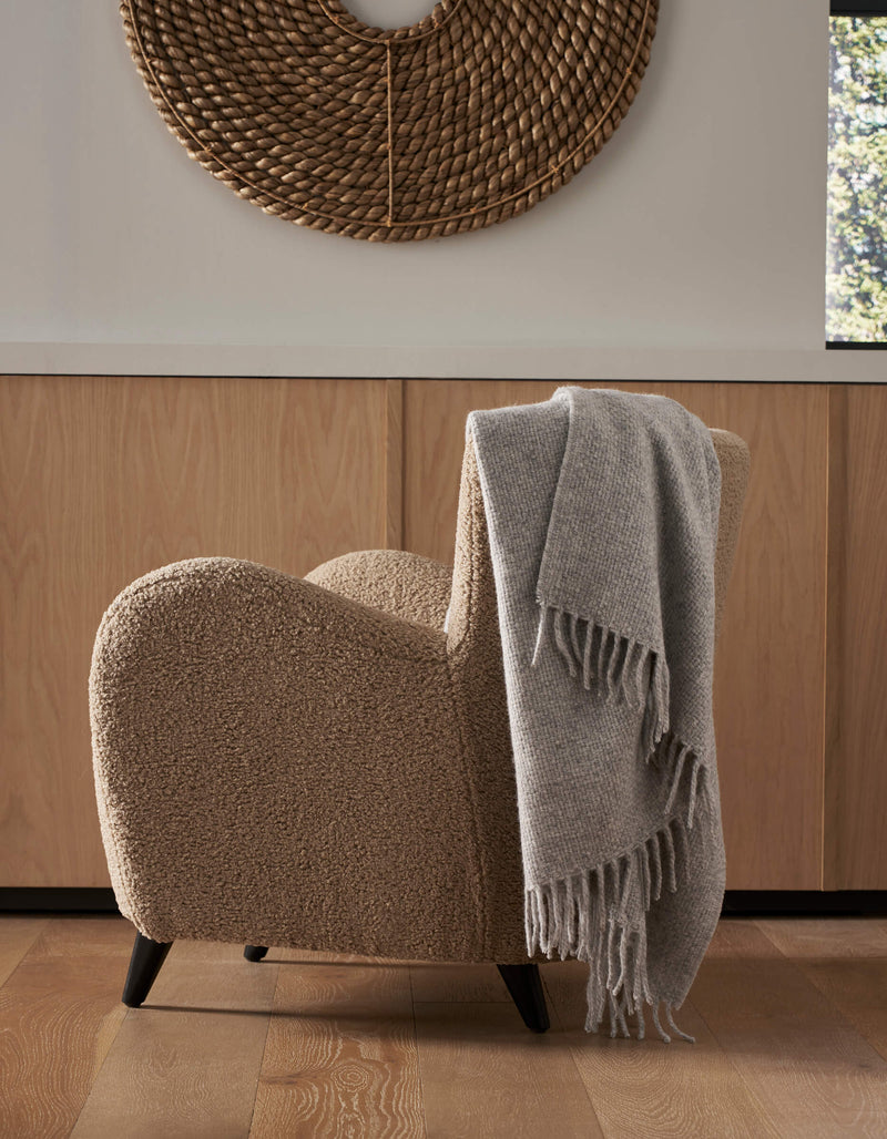 A cozy beige armchair with the Cozy Earth Lucia Throw in soft gray draped over its side sits on a wood floor. Behind it are light wooden cabinets and a round woven wall decor, with sunlight streaming through a nearby window.