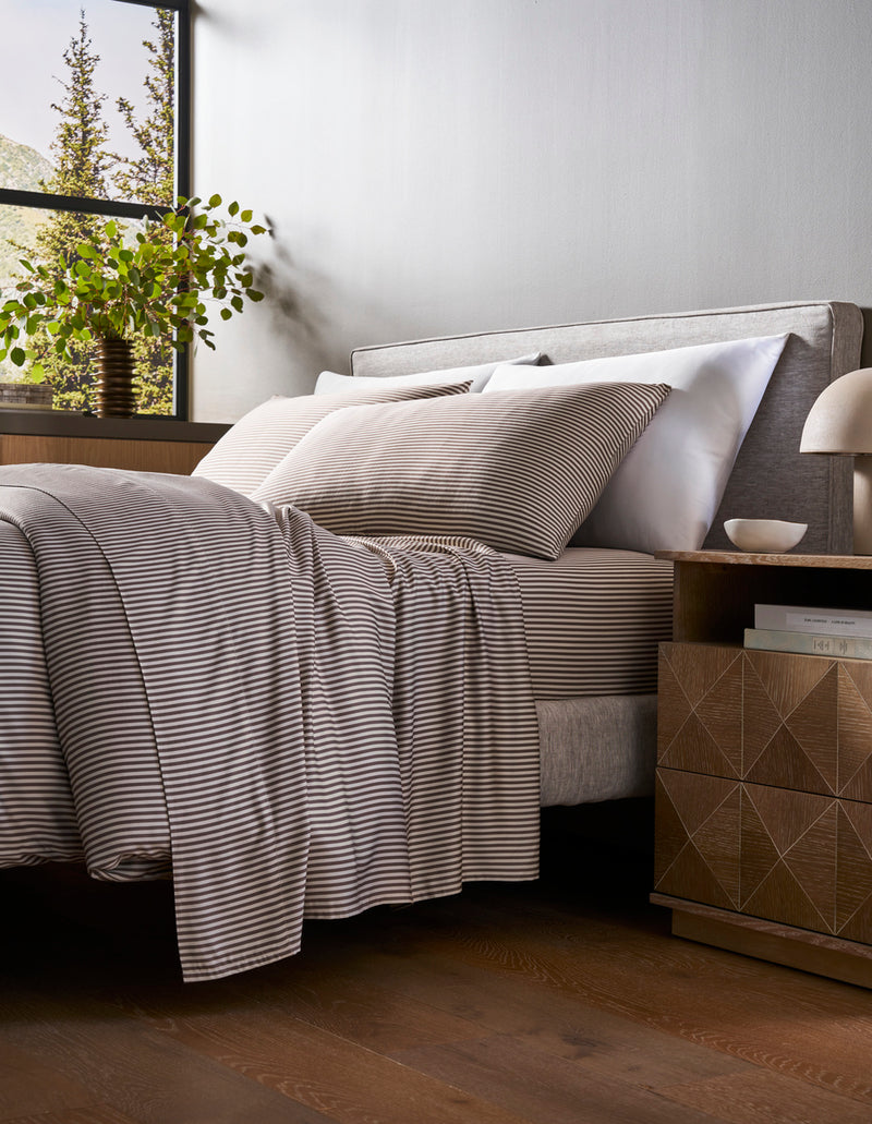 A bed with a Cozy Earth Bamboo Duvet Cover in gray and white stripes sits by a wooden nightstand topped with books and a lamp. Natural light streams in from a window where a potted plant rests on the sill, revealing trees outside.