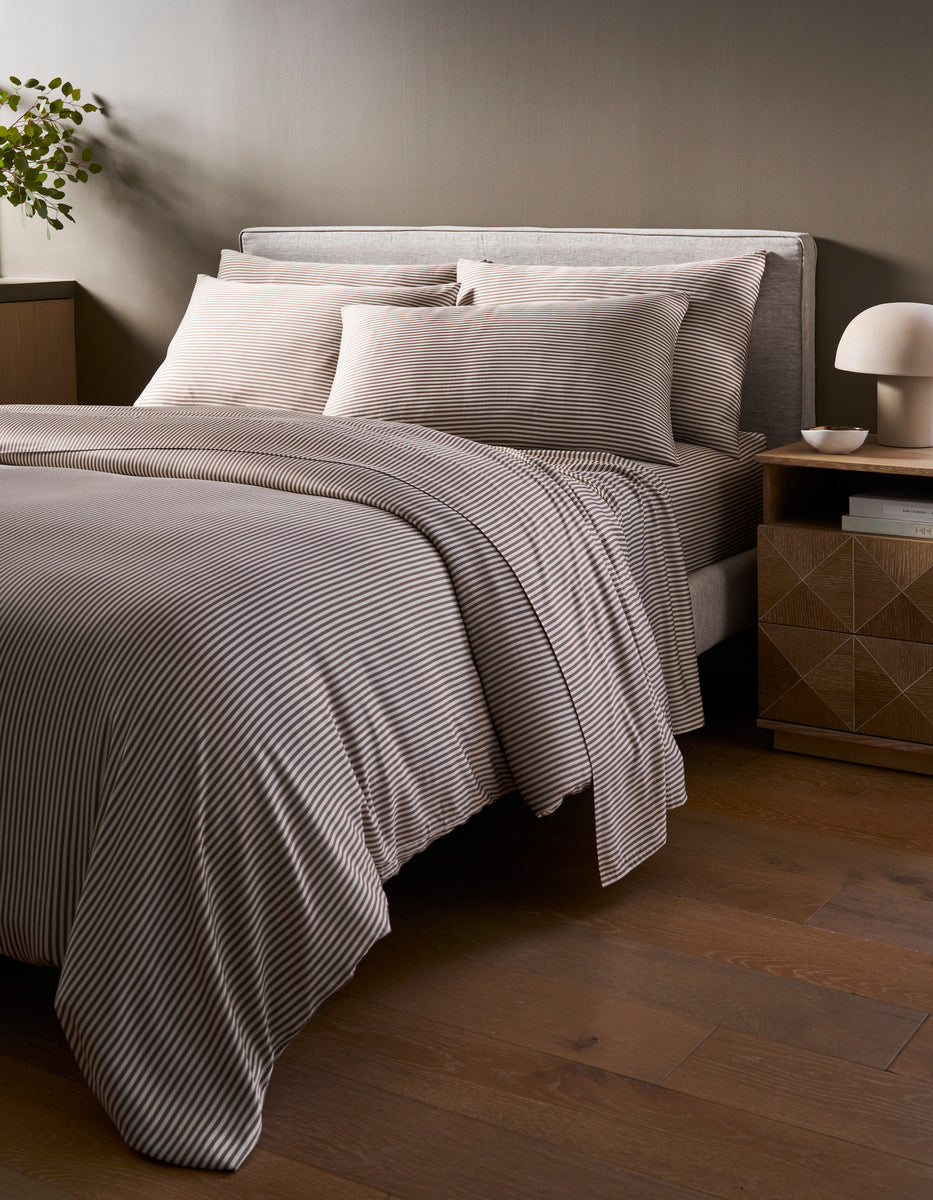 A neatly made bed with the Cozy Earth Bamboo Duvet Cover in gray and white stripes stands in a modern bedroom with wooden floors, a nightstand, a lamp, and a small plant in the corner.