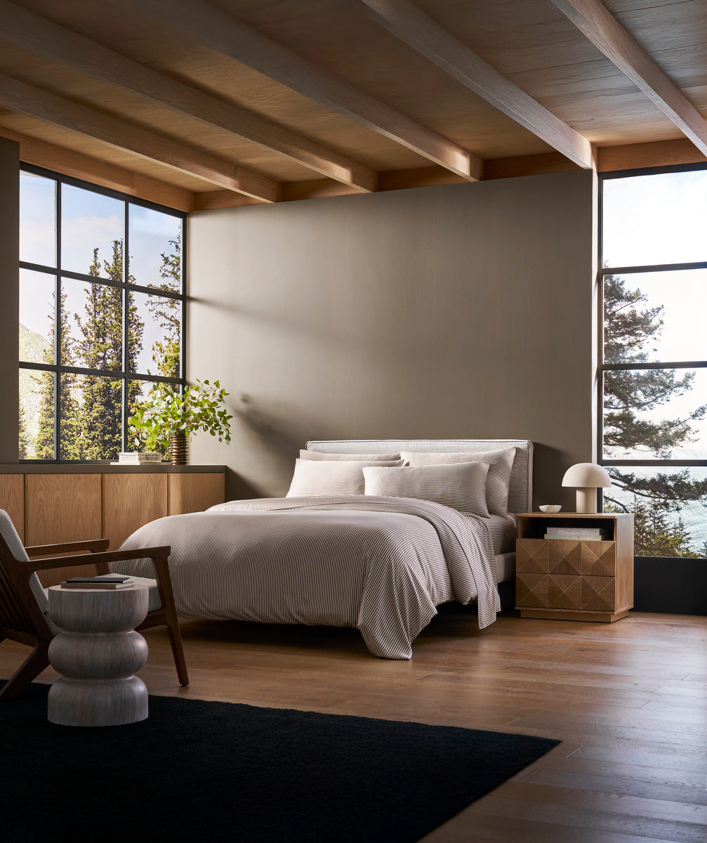 A modern bedroom with large windows, wood floors, a bed featuring the Cozy Earth Bamboo Duvet Cover in white, a wooden nightstand, lamp, black rug, a unique round stool, and views of trees outside.