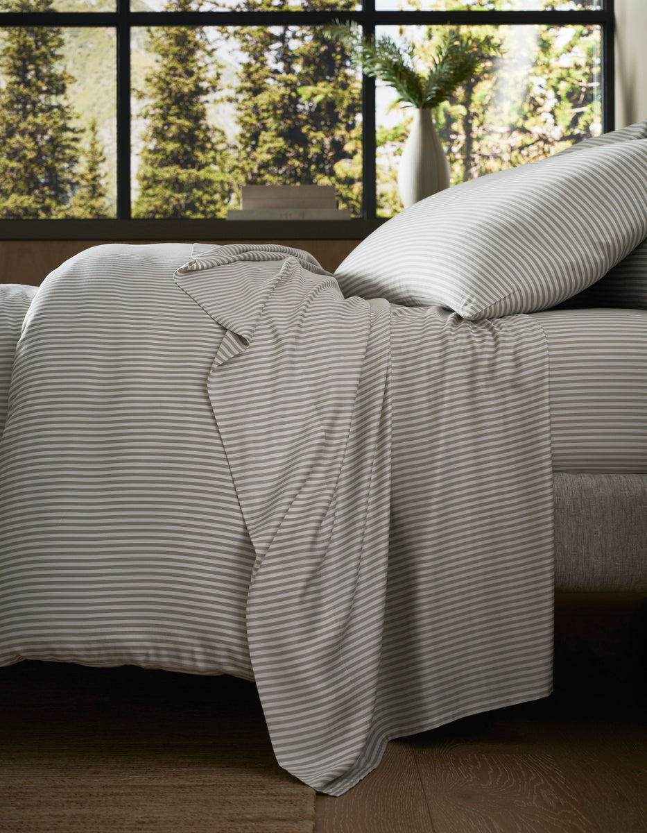 A neatly made bed with the Cozy Earth Bamboo Bedding Core Bundle in gray and white stripes sits by a large window, sunlight streaming in to illuminate a vase of green foliage on the windowsill and reveal views of trees outside.