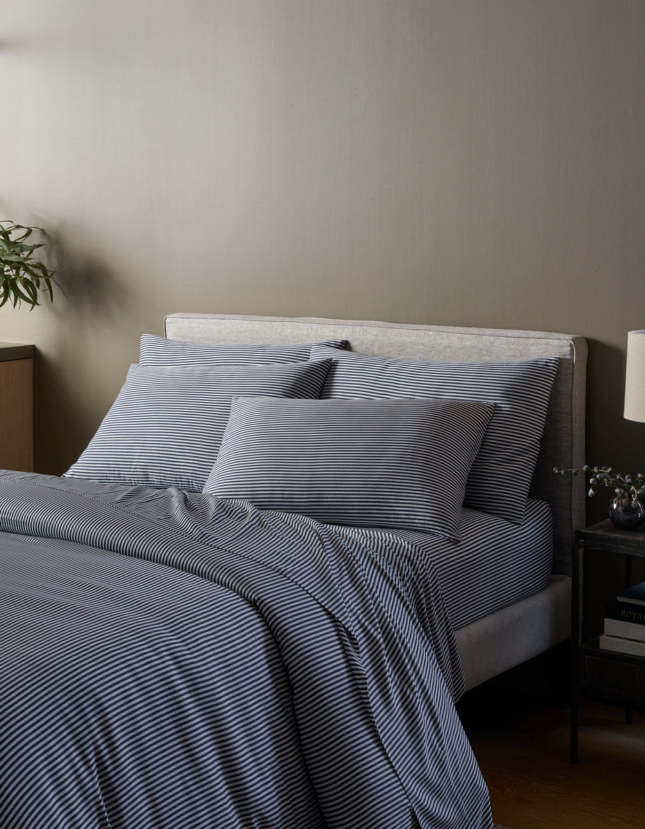 A neatly made bed features the Cozy Earth Bamboo Duvet Cover in gray and white stripes, complemented by matching pillows and a comforter. Against a beige wall, it’s flanked by a nightstand with a lamp, vase, and a plant in the corner.
