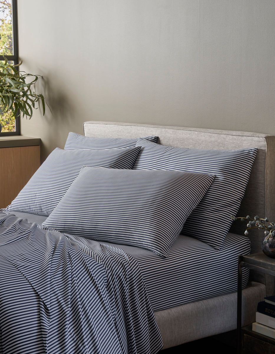 A neatly made bed with navy and white striped sheets and Cozy Earth Bamboo Pillowcases sits by a sunlit window. A green plant is on the cabinet, and a stack of books rests on the bedside table.