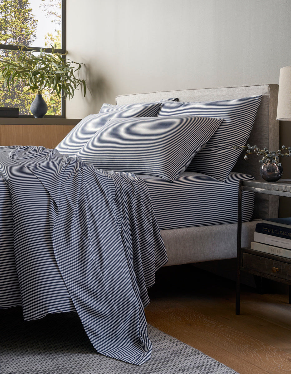 A modern bedroom features a neatly made bed with Cozy Earth’s Bamboo Bedding Core Bundle in black and white stripes, accompanied by a small nightstand holding books, a lamp, and a vase of flowers under soft lighting.