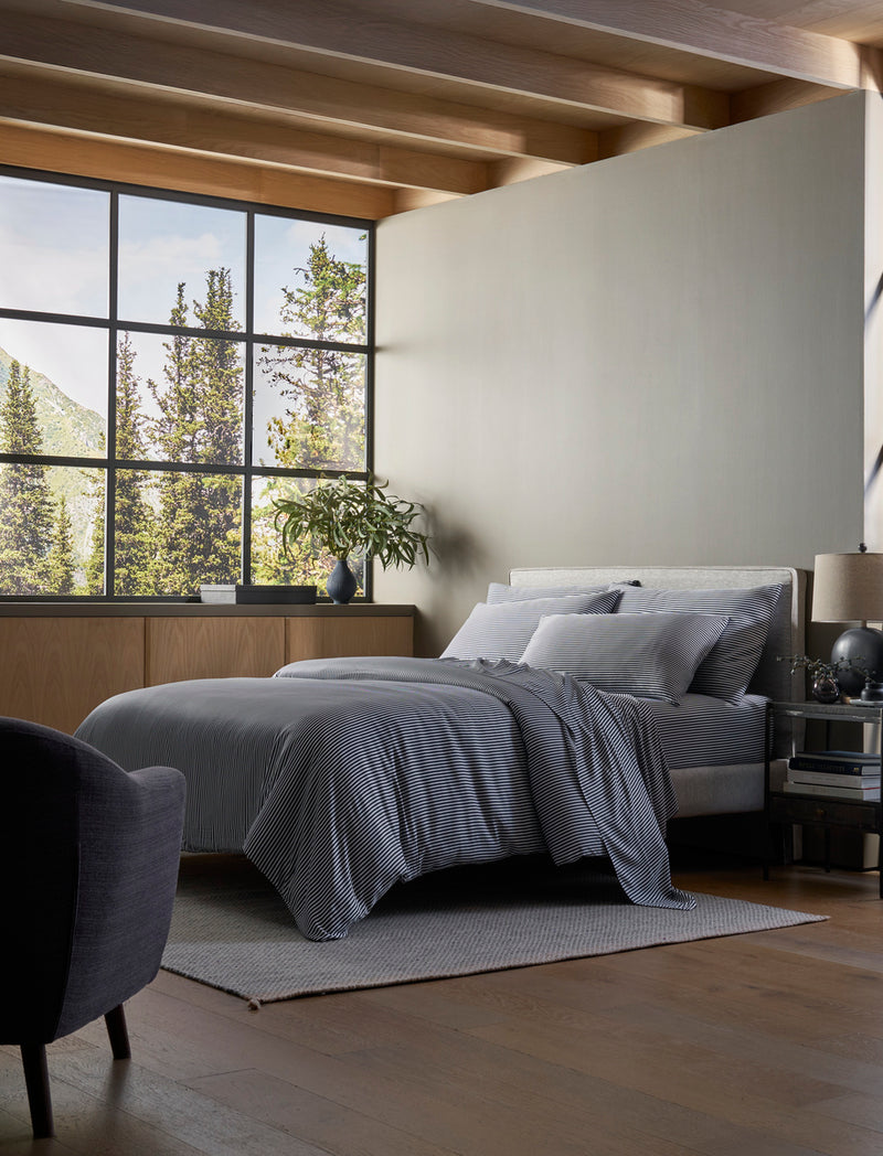 A modern bedroom with a large window, gray armchair, light rug, and wooden floors features a neatly made bed with Cozy Earth's Bamboo Duvet Cover. Natural light fills the space, with plants and decor displayed on the dresser.