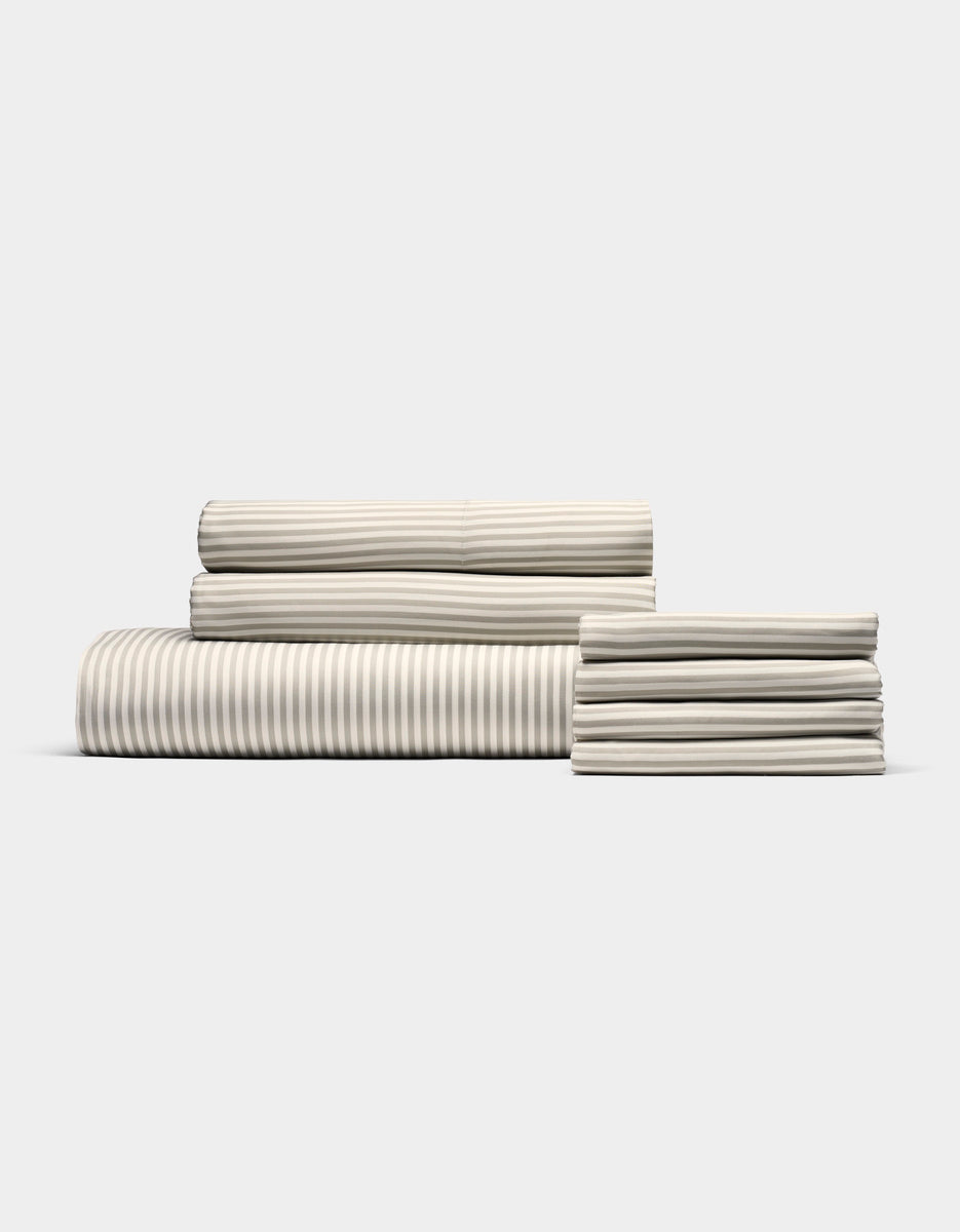 The Cozy Earth Bamboo Bedding Core Bundle features neatly folded cream-colored sheets with subtle light gray stripes, arranged on a plain white background.