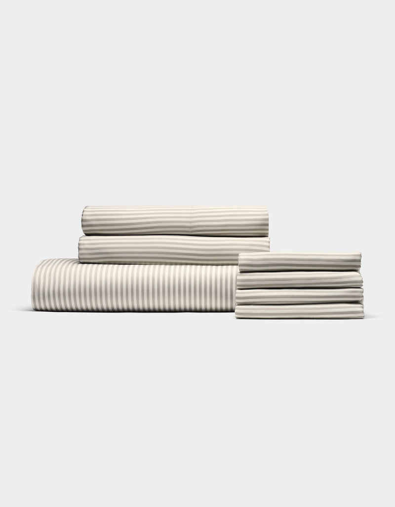 The Cozy Earth Bamboo Bedding Core Bundle features neatly folded cream-colored sheets with subtle light gray stripes, arranged on a plain white background.