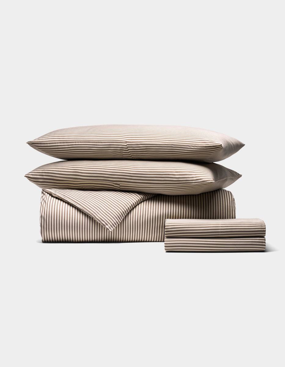 The Bamboo Bedding Deluxe Bundle by Cozy Earth features a neatly stacked beige and white striped set with two pillows, a folded comforter, and a folded sheet arranged on a plain light background.